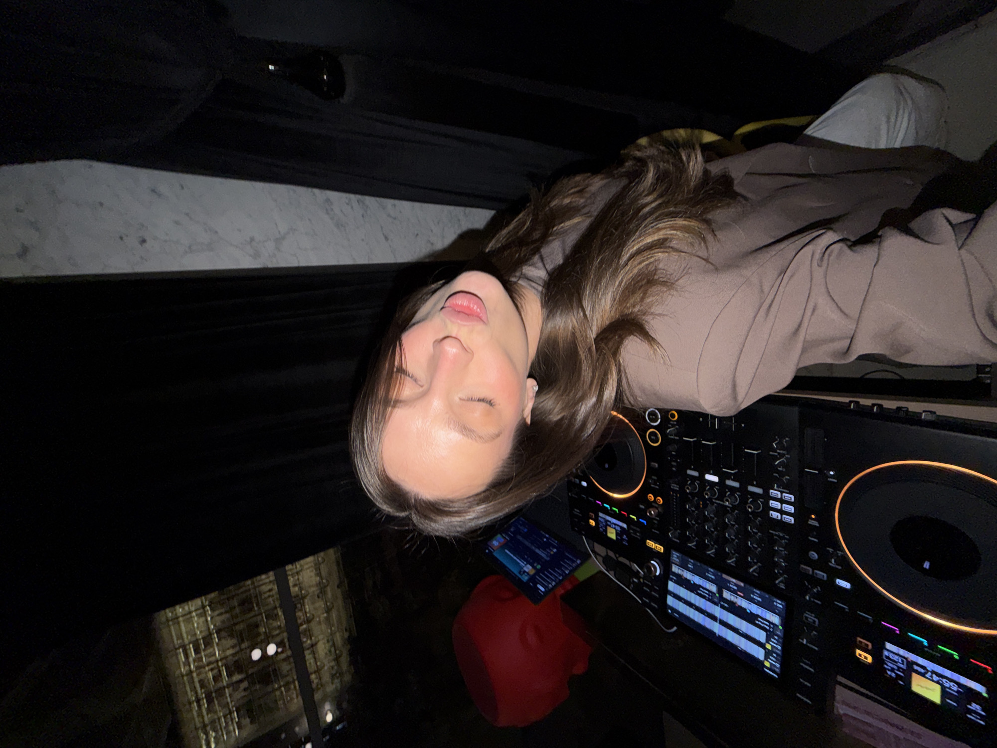 A woman lies with her eyes closed next to a DJ mixing console. She wears a light brown jacket and gray pants. The console is black and has glowing orange accents. A red mannequin head rests on a dark surface with a tablet. Outside the window is an illuminated building. This scene is likely taking place in Budva, Montenegro.