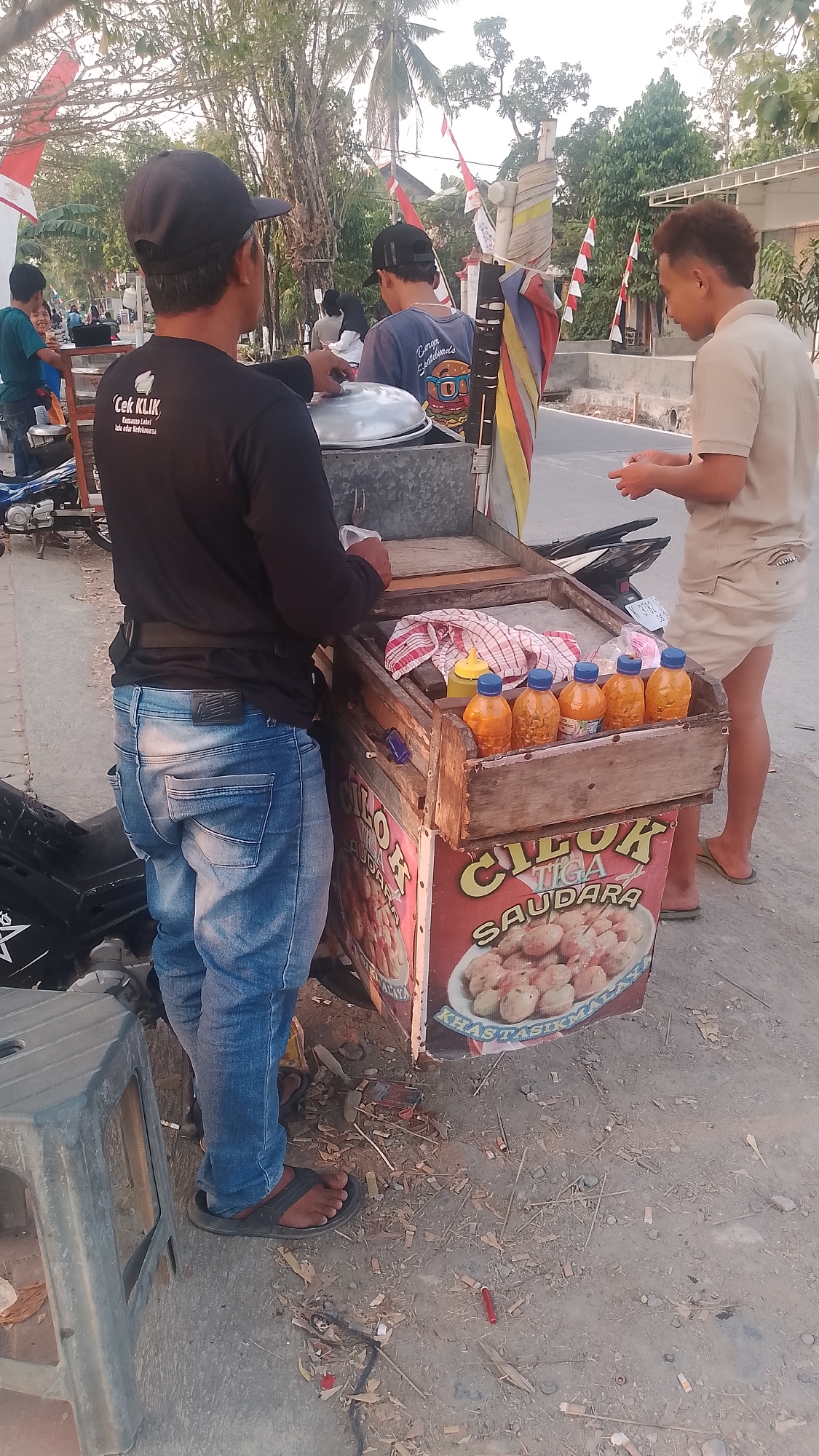 An outdoor, eye-level shot captures a street food vendor in Karanglangu, Indonesia, preparing food. The foreground is dominated by the vendor, a male wearing a black t-shirt with white text reading "Cok Klik," a black baseball cap, and blue jeans. He is standing with his back to the camera, attending to a metal pot on a cart. To his right, another male, dressed in a tan polo shirt and tan shorts, is seen from the side, holding a small white object, possibly food or change, with both hands as he interacts with the vendor.
The food cart, constructed of wood, features a prominent sign with red and yellow lettering that reads "CILOK TIGA SAUDARA" and displays an image of the food for sale. Five clear plastic bottles filled with an orange liquid, likely a sauce, are neatly arranged on top of the cart. Next to these bottles rests a folded red and white checkered cloth. A motorcycle is parked behind the cart.
The background reveals a street with a few more people, some walking, and a sidewalk. Trees with palm fronds line the street, and Indonesian flags, red and white, are visible, suggesting a local celebration or national holiday. The lighting indicates it is late afternoon or early evening, with warm sunlight casting long shadows. The ground is unpaved and scattered with debris, including dried leaves and small sticks.