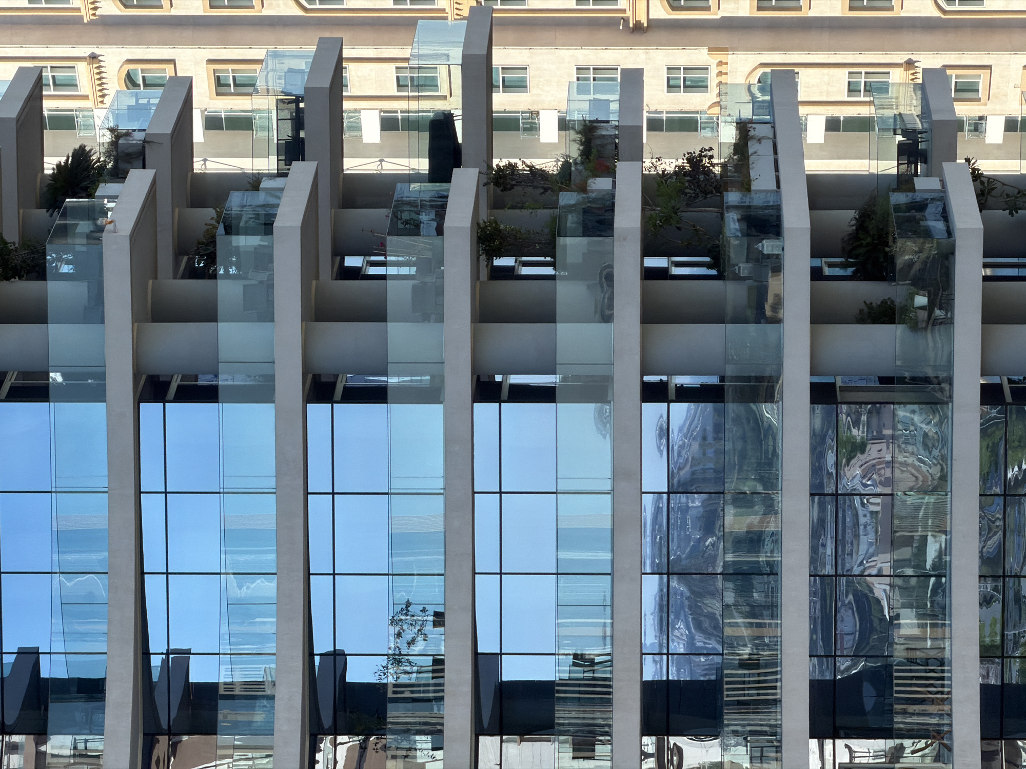 This is a detailed, forensic, and matter-of-fact description of an outdoor urban scene, focusing on architectural details. The image displays a modern building with multiple levels and balconies. The facade is dominated by large windows with a blue, reflective tint, suggesting the presence of a sky or a surrounding environment that is being mirrored. These windows are divided into a grid pattern.
Interspersed between the rows of windows are horizontal concrete structures, forming balconies and floor levels. These balconies are enclosed by transparent glass railings, some of which have a frosted appearance. Small potted plants and greenery are visible on some of these balconies, adding a touch of nature to the urban architecture. In some balconies, outdoor furniture, such as chairs and tables, can be discerned.
The building's structure is supported by substantial concrete columns. To the right of the main building, another structure is visible, appearing to be an adjacent building with a lighter colored facade, possibly beige or cream. This neighboring building also features multiple windows with a simpler design and arched tops on some levels. The overall impression is one of contemporary urban development, characterized by reflective surfaces and geometric forms. No people or animals are visible in the image.