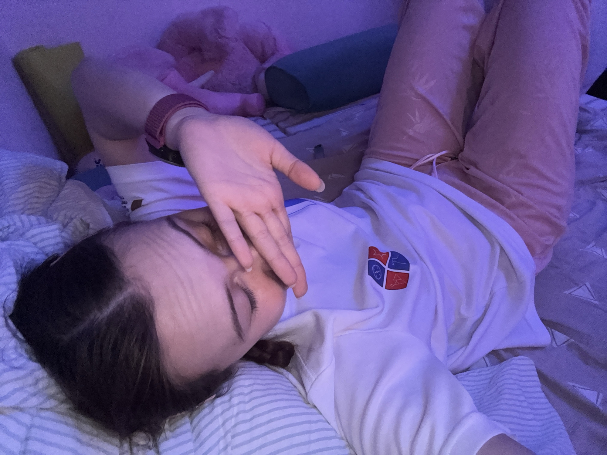 A person lies in bed, their face obscured by their hand. They wear a white sweatshirt with a logo on the chest and pink pants. The bed has striped sheets and a pillow. A stuffed animal and cylindrical pillow are visible in the background. The location is not discernible from the image.