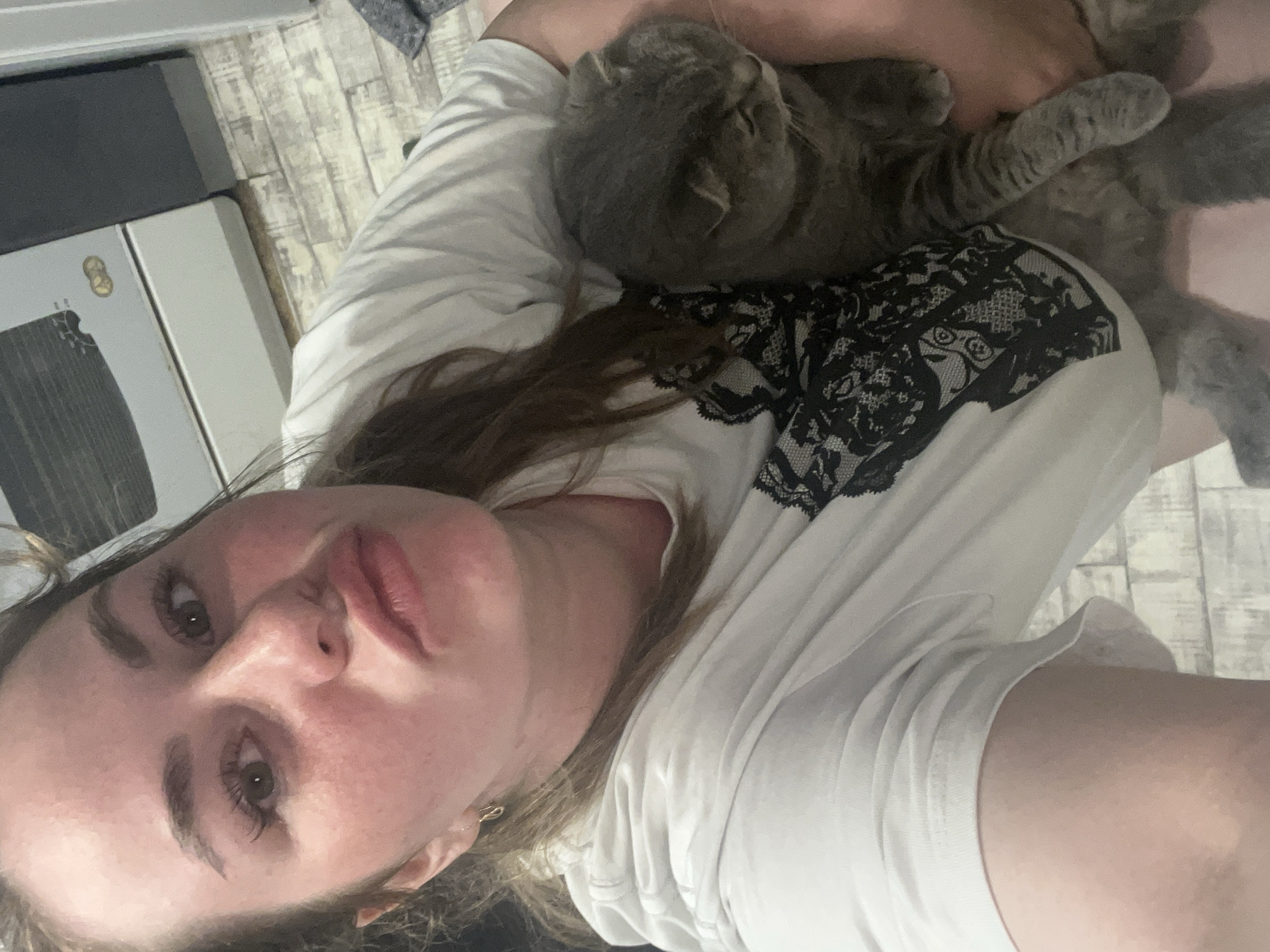 A close-up selfie captures a woman and a grey cat in an indoor setting in Chaikovsky, Russia. The woman, positioned at the bottom-left of the frame, looks directly into the camera with a neutral to slightly pouting expression. She has long brown hair, dark eyes, and a noticeable nose and lips. She is wearing a white t-shirt featuring an elaborate black lace pattern across the chest and shoulders. A small, simple gold earring is visible on her left ear.
Nestled comfortably on the woman's chest and shoulder area is a grey cat, likely a domestic short-haired breed, possibly with tabby patterns on its legs. The cat's head is turned away from the camera, and it appears relaxed and settled.
The background features a light-colored floor with a distinctive wood-grain or laminate pattern. To the left of the frame, a white household appliance, possibly a washing machine or similar unit, is partially visible. It has a dark top panel and a small, circular, gold-colored emblem or logo on its front. No discernible text is visible on the emblem or elsewhere in the image. The lighting suggests an indoor environment, typical of a home, depicting a casual moment of companionship between the woman and her pet.