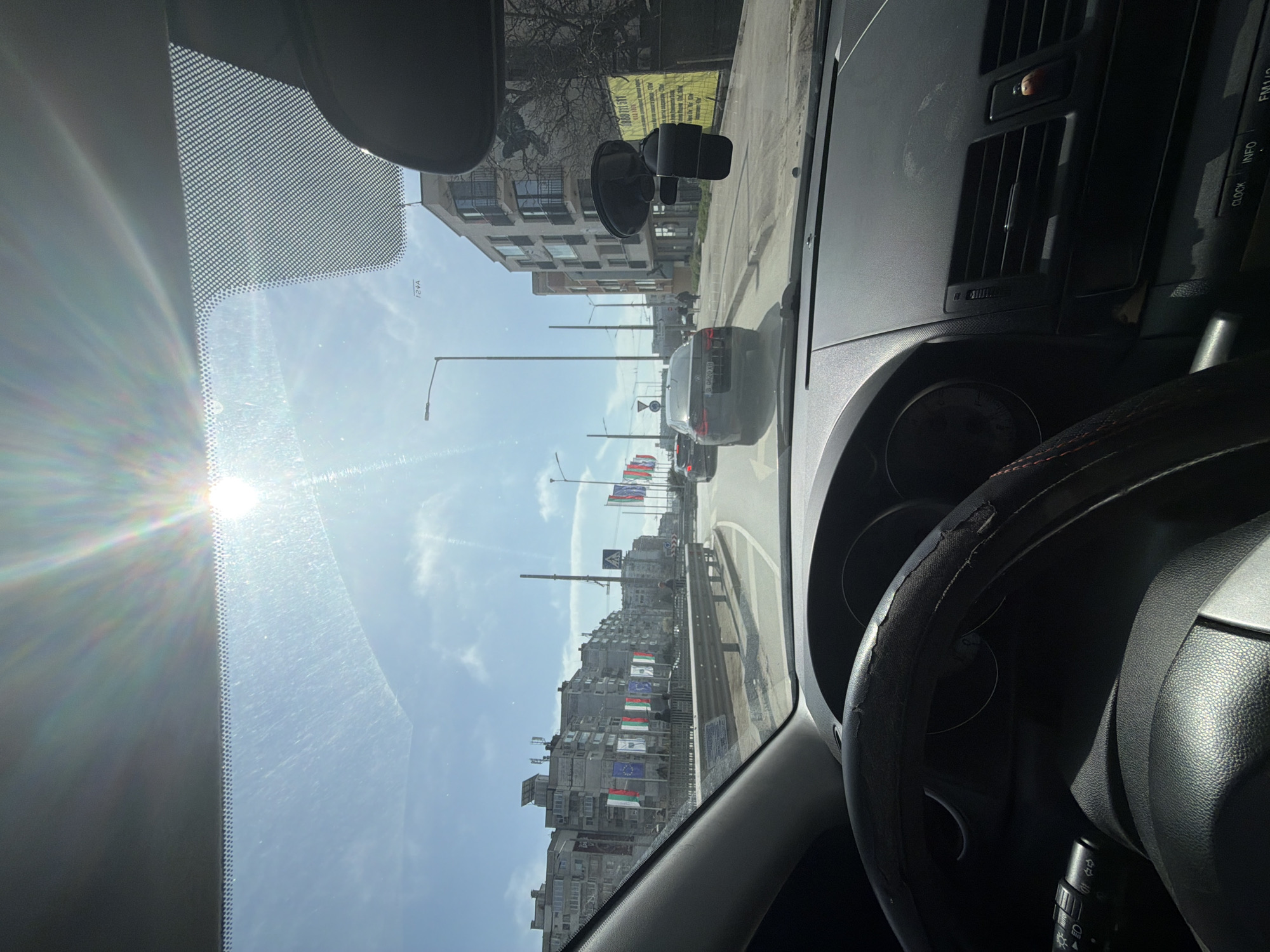 View from inside a car driving on bulevard "Vladislav Varnenchik" in Varna, Bulgaria. In the foreground is the car's dashboard, steering wheel, and windshield. Through the windshield, the street is visible with cars, buildings decorated with flags, and streetlights. The sun is shining brightly.