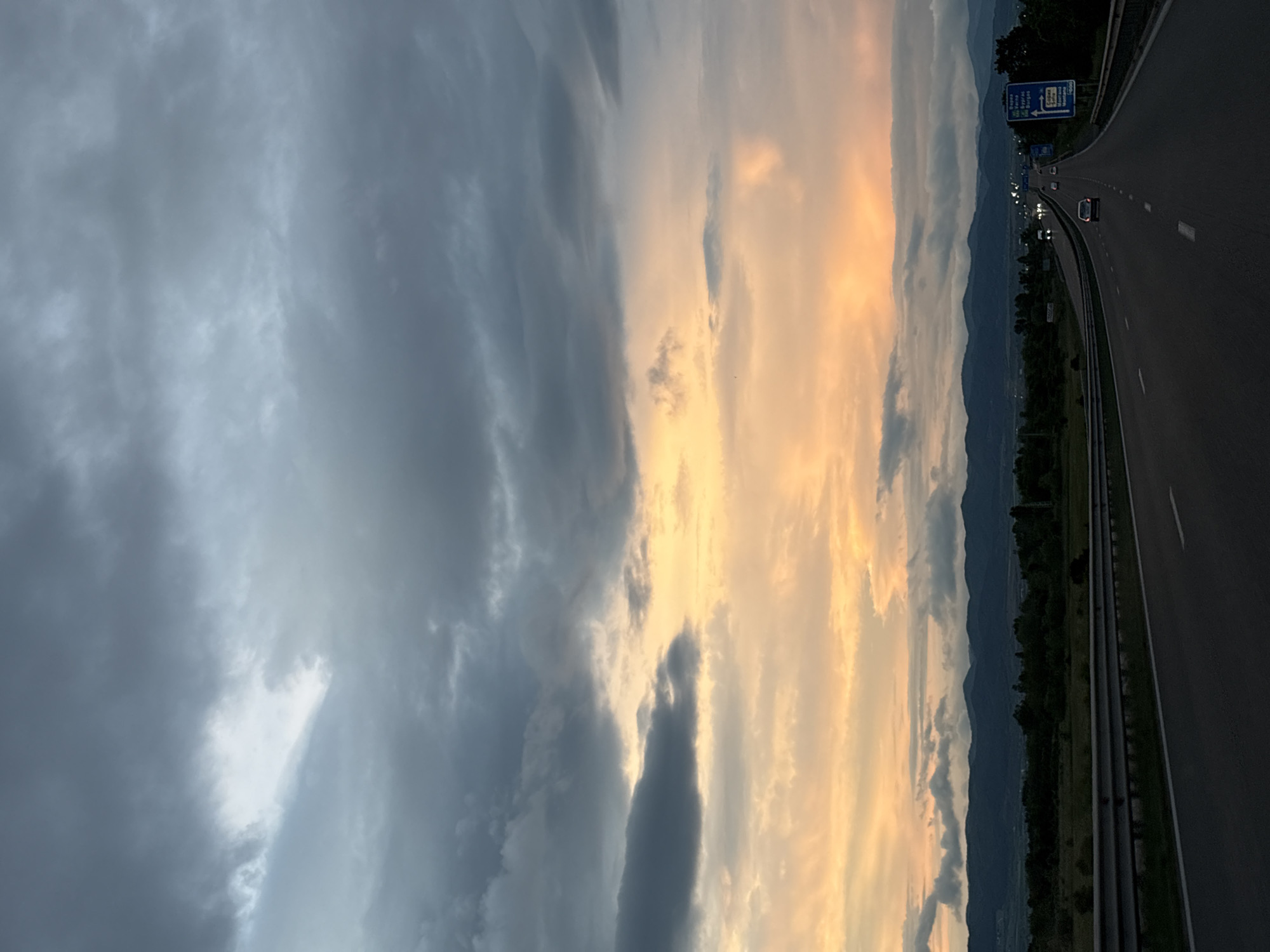 The image depicts a view from a highway near Sofia, Bulgaria. The road stretches into the distance with mountains on the horizon. A road sign is visible on the right. The sky is dominated by dramatic cloud formations, with a blend of dark storm clouds on one side and lighter, sunset-colored clouds on the other. There are a few cars on the road.