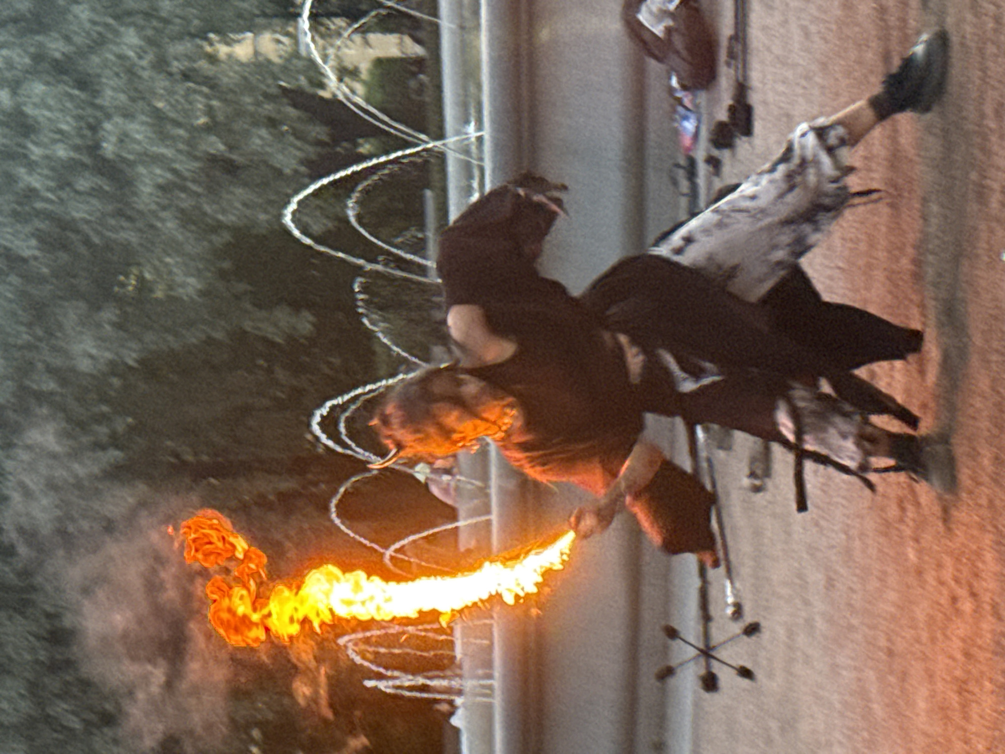 A person is performing with fire in front of a fountain in St. Petersburg, Russia. The performer, wearing dark clothing and patterned pants, is holding a flaming object that is shooting a plume of fire upwards and to the left. Their hair is dark and appears to have horns attached. In the background, a fountain is spraying water. The ground is a sandy or gravelly surface. There are other objects on the ground, including what appear to be props for the performance.