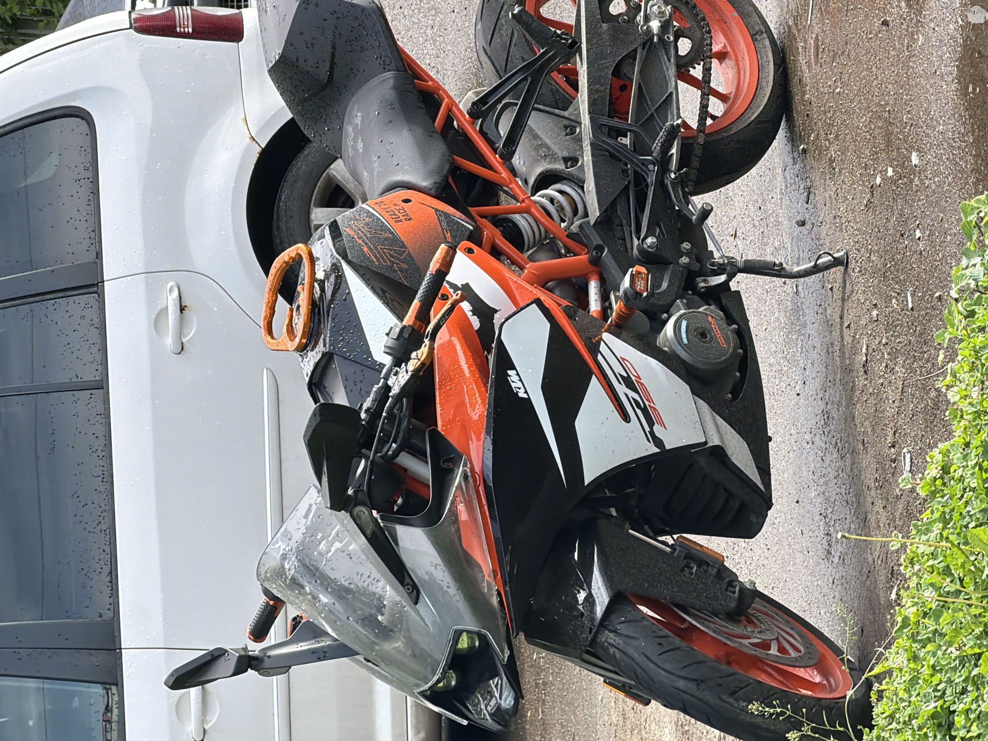 On an undetermined street in the city of Unknown, Unknown, a KTM motorcycle is leaning against a white vehicle, apparently damaged. The motorcycle displays orange and black components, while the vehicle is a light-duty van or passenger vehicle. The scene is outdoors, with asphalt pavement visible.