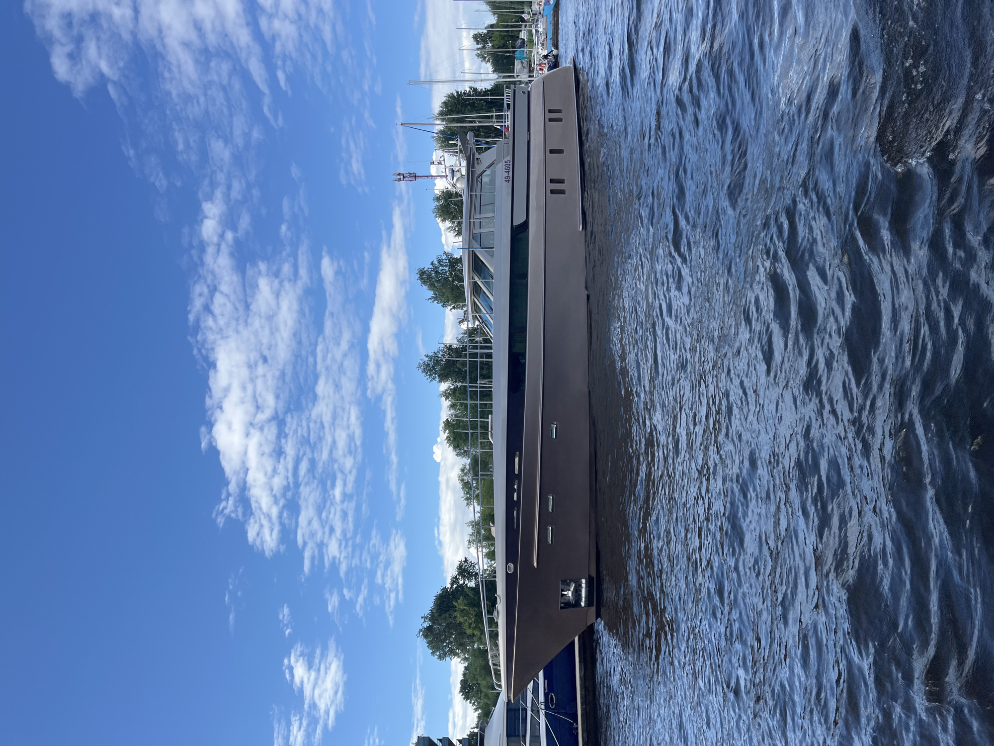 The image shows a large, modern yacht docked on a body of water, likely a river or canal, with Saint Petersburg's characteristic skyline visible in the background. The yacht has a sleek, contemporary design with a light-colored hull and large windows. It appears to be a pleasure craft, possibly for leisure cruises or day trips.
The scene is set on a bright, sunny day with a clear blue sky dotted with fluffy white clouds. The water is calm, with gentle ripples reflecting the sunlight, suggesting a pleasant, possibly warm, atmosphere. The surrounding area appears to be a marina or waterfront promenade, with lush green trees lining the shore and other boats, including sailboats, visible in the distance.
There is no visible text in the image, except for the boat's registration number, "49-4605", which is clearly printed on the side of the yacht. The overall mood is serene and peaceful, conveying a sense of relaxation and enjoyment often associated with boating and waterfront activities. The presence of other vessels and the verdant landscape suggest a well-developed urban waterfront area, typical of a major European city like Saint Petersburg.