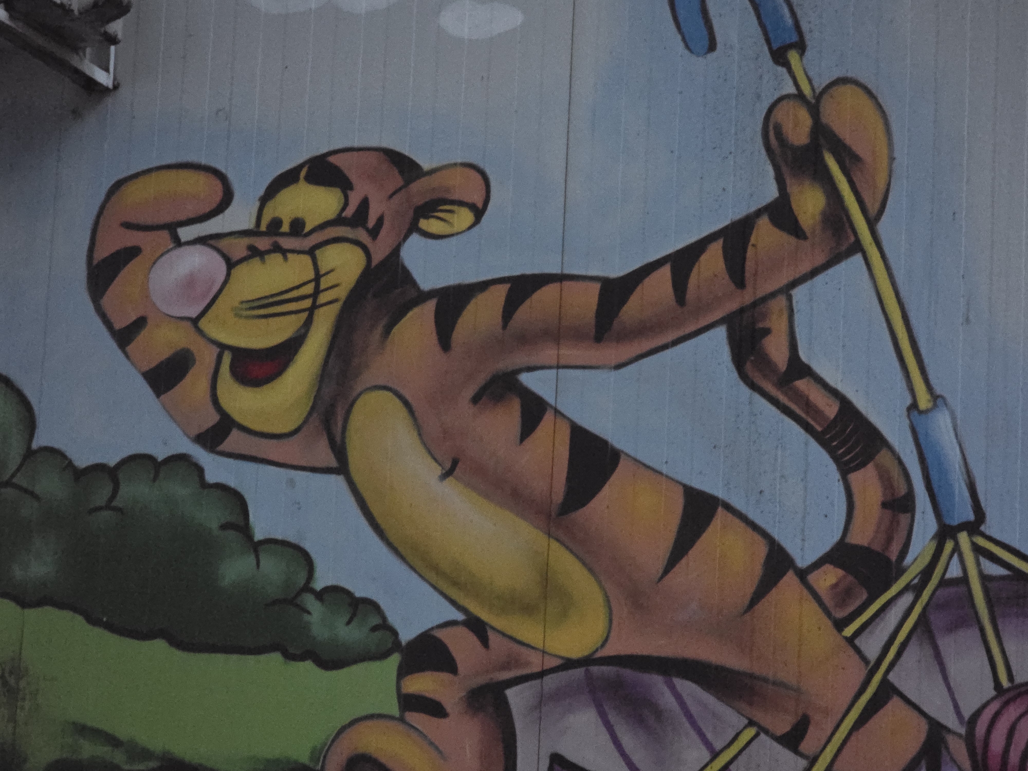 This media file features a colorful, cartoon-style mural depicting a tiger character, strongly resembling Tigger from Winnie the Pooh. The tiger is the central subject, rendered in orange with black stripes, a yellow muzzle and belly, and a pink nose. It wears a wide, cheerful smile, suggesting joy and energy.
The tiger appears to be in motion, perhaps swinging or hanging from a playground-style ride. Its body is dynamically posed, leaning back with one arm bent and raised near its head, and the other arm extended to grip the yellow bars of a swing or similar apparatus. The swing structure includes blue connectors and hints of purple and white elements at the bottom, possibly indicating a seat or part of the ride.
In the background, a simplified outdoor scene is depicted. The lower left features dark green, cloud-like bushes or foliage, while the upper background is a clear, light blue sky with faint, white cloud shapes. The overall lighting is bright and even, suggesting daytime. The image has a textured quality, indicating it is likely a painting or mural on a wall, with subtle vertical lines running across the surface. No text is visible in the image.
