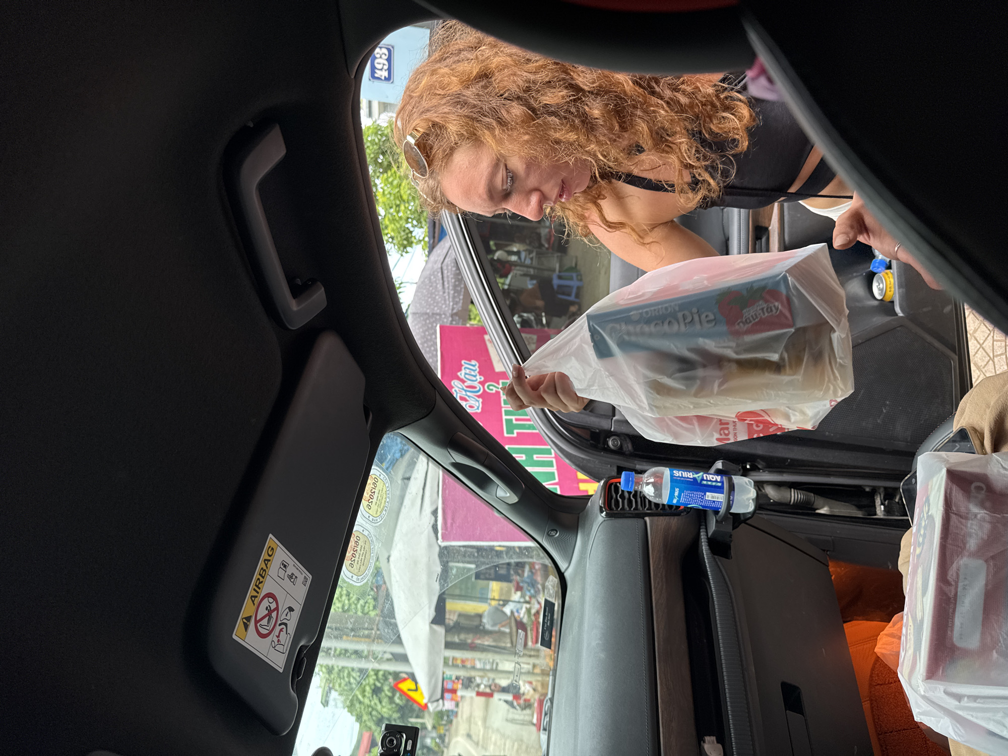 The image is taken from inside a car, looking out towards the right side. A woman with curly, red hair, wearing a black tank top and sunglasses pushed up on her head, is in the passenger seat. She is holding a clear plastic bag filled with food items, appearing to be reaching for something or handing it over. In the background, through the car window, a street scene is visible in Bắc Yên, Vietnam. There are buildings, street vendors with umbrellas, and people on the street. A yellow triangular traffic sign is also visible. Inside the car, a bottle of Aquarius water is placed in a cup holder on the door. Another plastic bag containing food items is on the floor in the foreground. The car's interior includes an airbag warning sticker on the sun visor and a grab handle on the ceiling. The overall impression is a snapshot of a transaction or a pause during a journey on a street in Bắc Yên.