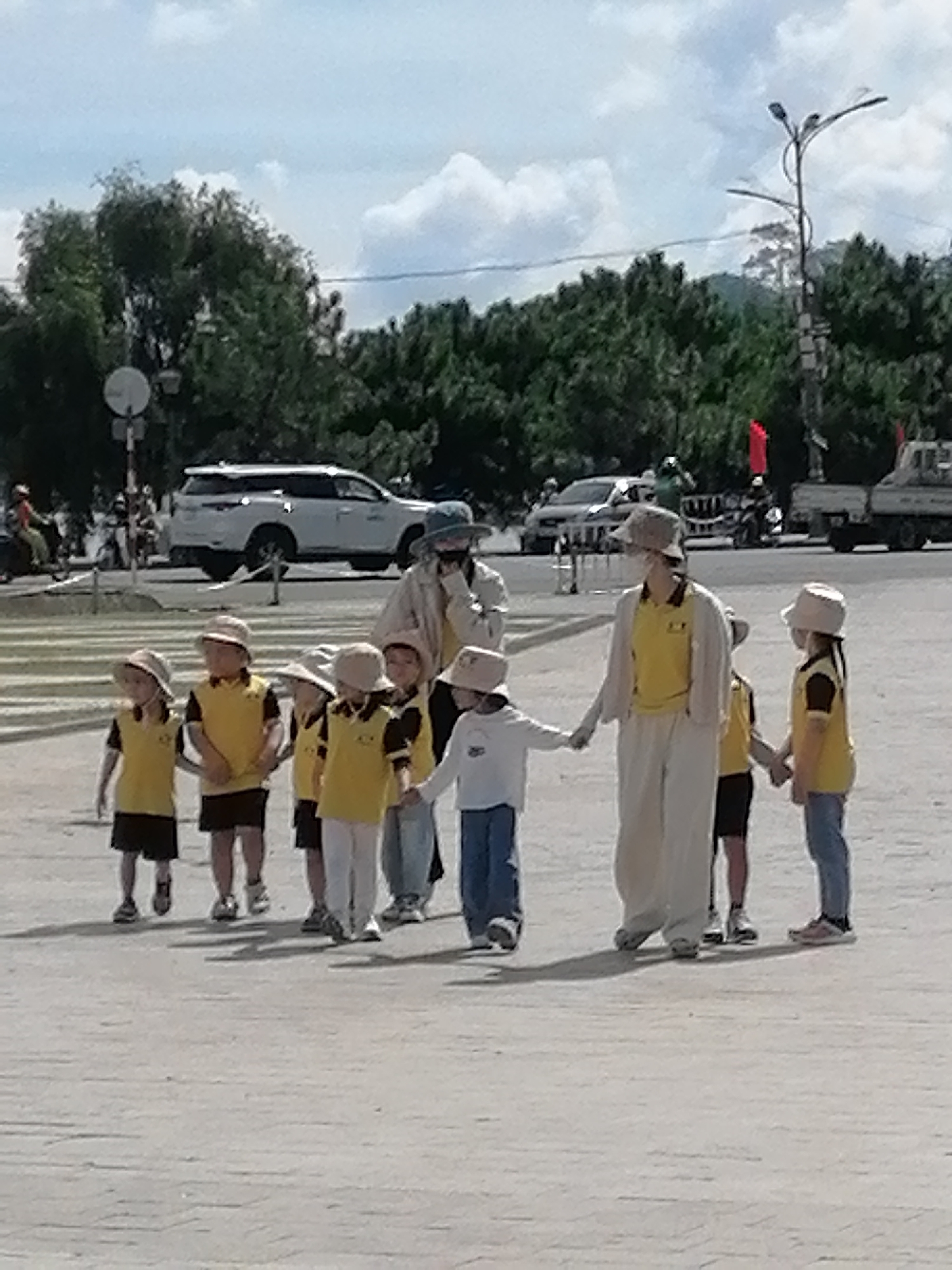 The image depicts a group of children, appearing to be of primary school age, walking in a line holding hands. They are wearing matching yellow shirts with dark trim and dark shorts or skirts, along with light-colored hats. Two adults, presumably teachers or chaperones, are accompanying them. One adult, dressed in a light-colored outfit with a yellow shirt, is at the front of the line of children, and another adult, wearing a light-colored jacket and a hat, is slightly behind and to the right of the children.
The setting appears to be a public space, possibly a pedestrian crossing or a plaza, paved with light-colored tiles that exhibit a brick-like pattern. In the background, several vehicles are visible, including a white SUV, a white truck, and motorcycles. A lamppost with multiple lights is also present in the background, along with trees and a hillside covered in dense greenery. The sky is partially cloudy. The overall scene suggests an outdoor activity or excursion involving children in a city environment.
Based on the visual cues, such as the architecture and the presence of vehicles commonly seen in Vietnam, combined with the typical urban landscape and vegetation, the location is likely Da Lat, Vietnam. Specifically, the scene might be near a tourist area or a school zone where pedestrian crossings are prevalent, given the presence of the vehicles and the paved walkway.