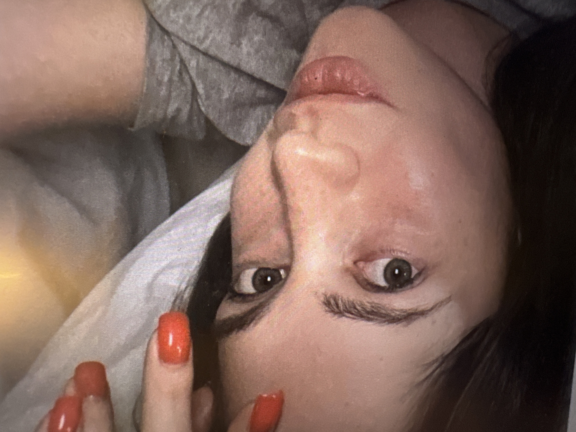 The image shows a close-up of a person's face, with their head tilted to the side and their eyes looking upwards. The person has dark hair and is wearing a grey garment. Their fingernails are painted bright red. The lighting suggests an indoor setting, possibly a bedroom or lounge area, with soft, diffused light and some highlights. There is no indication of the city of Murcia, Spain, or any other specific geographical location in the image itself. The scene appears to be a personal, intimate shot, possibly a selfie given the close-up perspective and the framing of the face.
