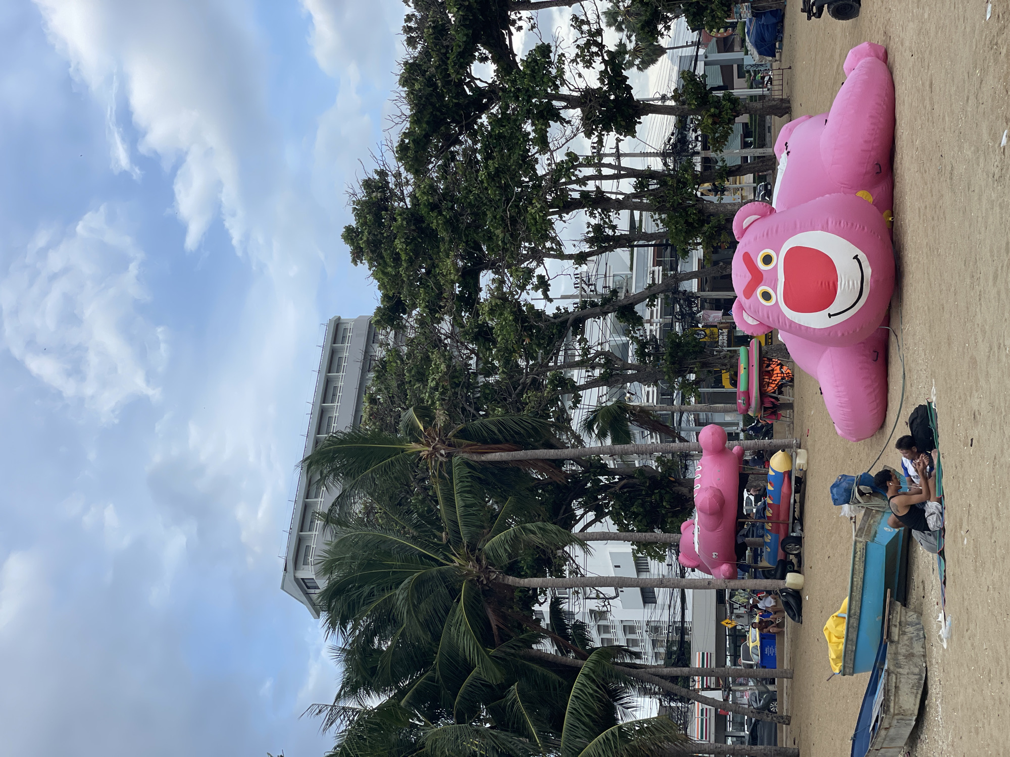 On a sandy beach in Pattaya City, Thailand, two individuals are lying on a mat in front of a blue boat. A large, pink inflatable bear, resembling Lotso from Toy Story, is deflated on the sand. In the background, a long, pink inflatable is propped up above a cart with inflatable tubes, possibly for water sports. Palm trees and other foliage are scattered across the scene, with a large building visible behind them. The sky is cloudy.