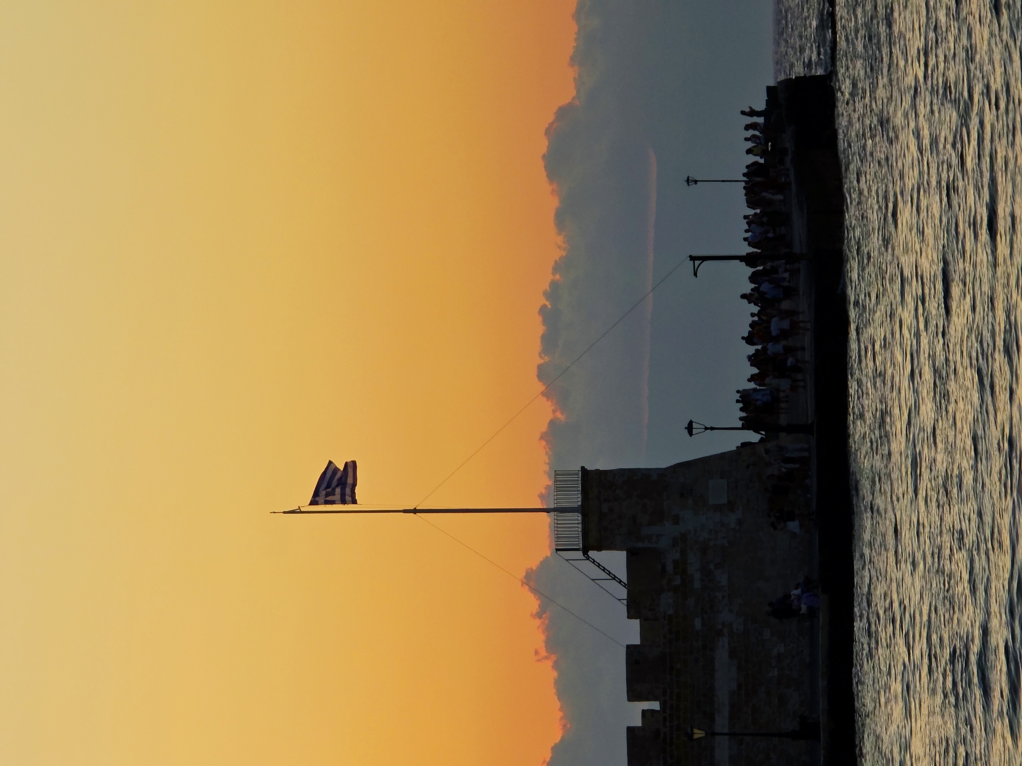 At sunset, a stone pier juts out into the sea in Chania, Greece. A Greek flag flies from a tall pole atop a fortified structure on the pier. Silhouetted figures, likely tourists, gather on the pier, some observing the flag and others facing the sea. The water is choppy, reflecting the warm orange hues of the sky. The scene is serene, capturing a moment of twilight in the harbor.