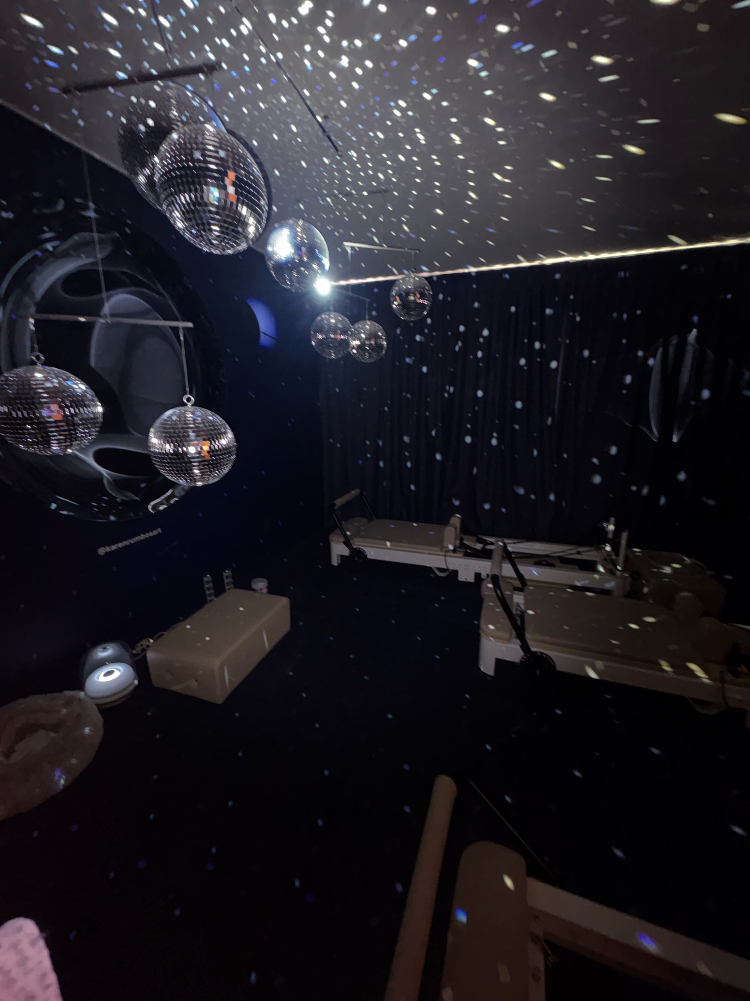 The image shows an indoor space decorated with disco balls and projected star-like lights, creating a celestial or starry night effect. Several disco balls are suspended from the ceiling, reflecting light and creating scattered patterns on the walls and ceiling. The room appears to be a studio or gym, as two Pilates reformers are visible on the right side of the image. In the foreground on the left, there is a light-colored, rectangular object, possibly a bolster or mat, and a round object that could be part of a yoga mat or cushion. The walls are dark blue, with some abstract circular or swirling patterns on the left side, and dark curtains with white speckles on the right, further enhancing the starry theme. The floor is dark and also reflects the projected lights. The overall impression is of a whimsical and energetic space, possibly designed for fitness classes or events with a fun, vibrant atmosphere. The location is not specified.