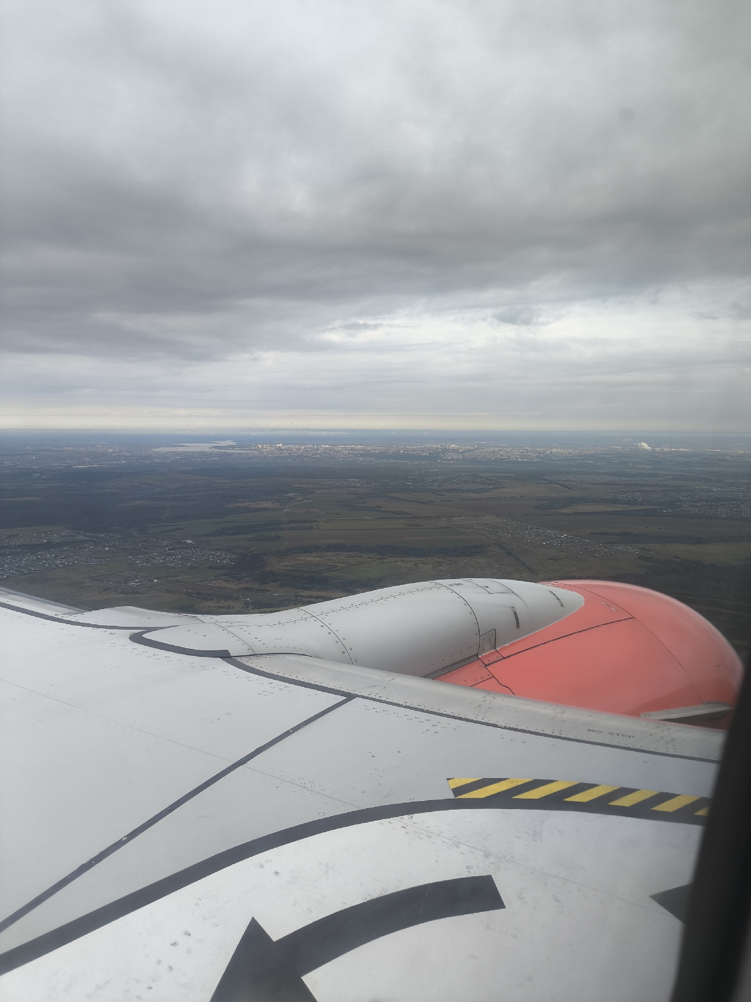 An aerial, eye-level shot captures a view from an airplane window looking out over a vast, sprawling landscape under a cloudy sky. In the foreground, the metallic surface of an airplane wing, painted white and grey, is visible, with visible rivets and dark grey stripes. A portion of a bright orange engine cowling is also present. The wing also features a black arrow marking and a yellow and black striped "NO STEP" warning decal. Beyond the wing, the landscape unfolds into a patchwork of green fields, dark forests, and scattered residential areas. In the distance, a dense urban area is visible, appearing as a collection of buildings and structures. The horizon is hazy, with the sky filled with thick, grey clouds, suggesting an overcast or potentially rainy day. No people or specific actions are discernible in the image.