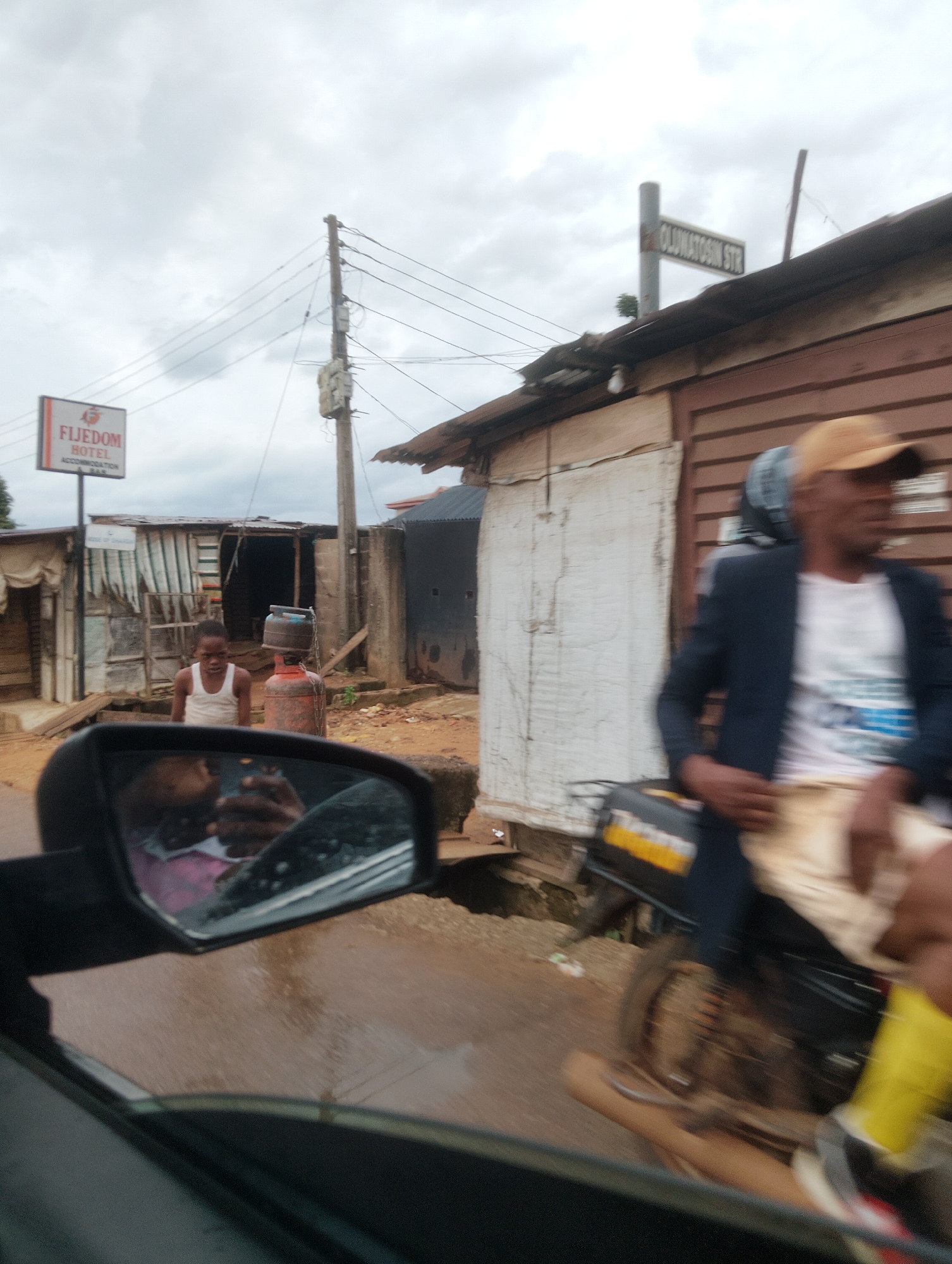 This image depicts a street scene in Iju, Nigeria, with a focus on a roadside setting. A young boy wearing a white tank top stands near a red gas cylinder. To the right, a person wearing a blue jacket, white t-shirt, and tan shorts is seated on a motorcycle, with their face blurred by motion. A street sign indicating "OLUWATOSIN STR" is visible in the upper right portion of the image, attached to a utility pole. Further left, a sign for "FIJEDOM HOTEL ACCOMMODATION BAR" is mounted on a pole. The background features dilapidated structures made of wood and corrugated metal. The ground appears to be dirt, and the sky is overcast. The scene is captured from within a vehicle, as evidenced by the reflection in a side mirror, which shows the interior of the vehicle and the upper bodies of two individuals.