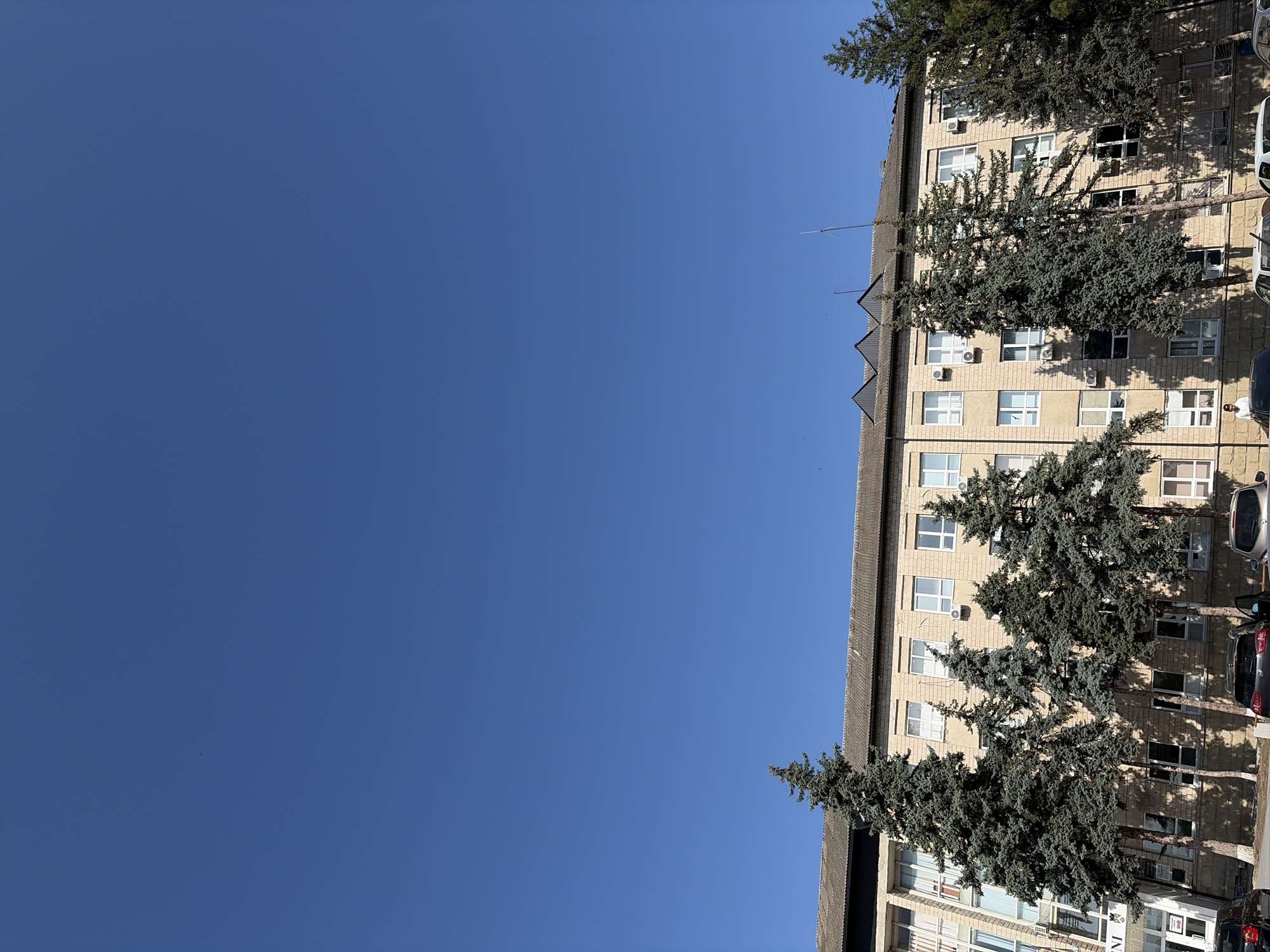 A low-angle, full-frame shot captures a light-colored, multi-story building with a grey, sloped roof, and a clear, deep blue sky. Tall evergreen trees, with their branches casting shadows on the building's facade, are spaced in front of the structure. The building features numerous rectangular windows, some with visible air conditioning units beneath them. The facade is constructed of light brown bricks. In the foreground, several cars are parked, with some doors open. A few individuals are visible near the parked vehicles and the building's entrance. The scene is set in the city of Criuleni, Moldova.