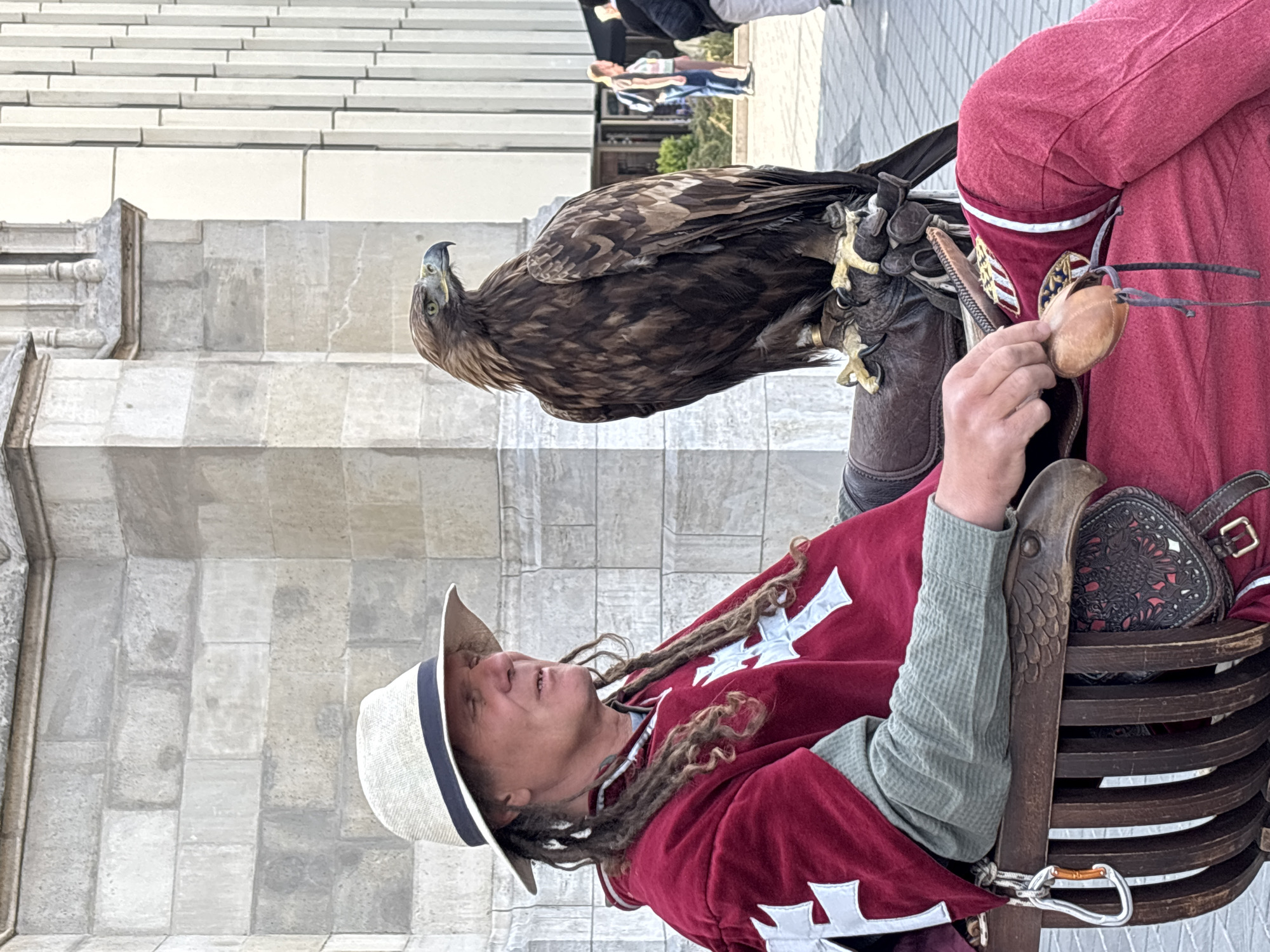 The image displays a close-up of a person dressed in historical-style attire, interacting with a large bird of prey. The person, who has dreadlocks and wears a light-colored straw hat, is seated in a dark wooden chair. They are dressed in a reddish-maroon tunic or cloak with several white cross symbols, resembling a medieval crusader's garb. Their left hand holds a small, light brown leather object, possibly a falconry hood or jess.
Perched on the person's right arm, which is gloved with a heavy leather gauntlet, is a large, brown bird with a hooked beak, appearing to be a Golden Eagle or a similar raptor. The bird is facing away from the viewer and looking upwards and slightly to its right. Its talons are visible gripping the gauntlet.
The setting appears to be outdoors, with a paved ground made of large, light-colored stone tiles. In the background, there is a large building or wall constructed from light-toned stone blocks, suggesting historical architecture. Other blurred figures of people are visible in the distant background, indicating a public area. The lighting is bright and natural, suggesting it is daytime under clear weather conditions. The scene evokes a public demonstration of falconry or an historical re-enactment event. No visible text is present in the image.
