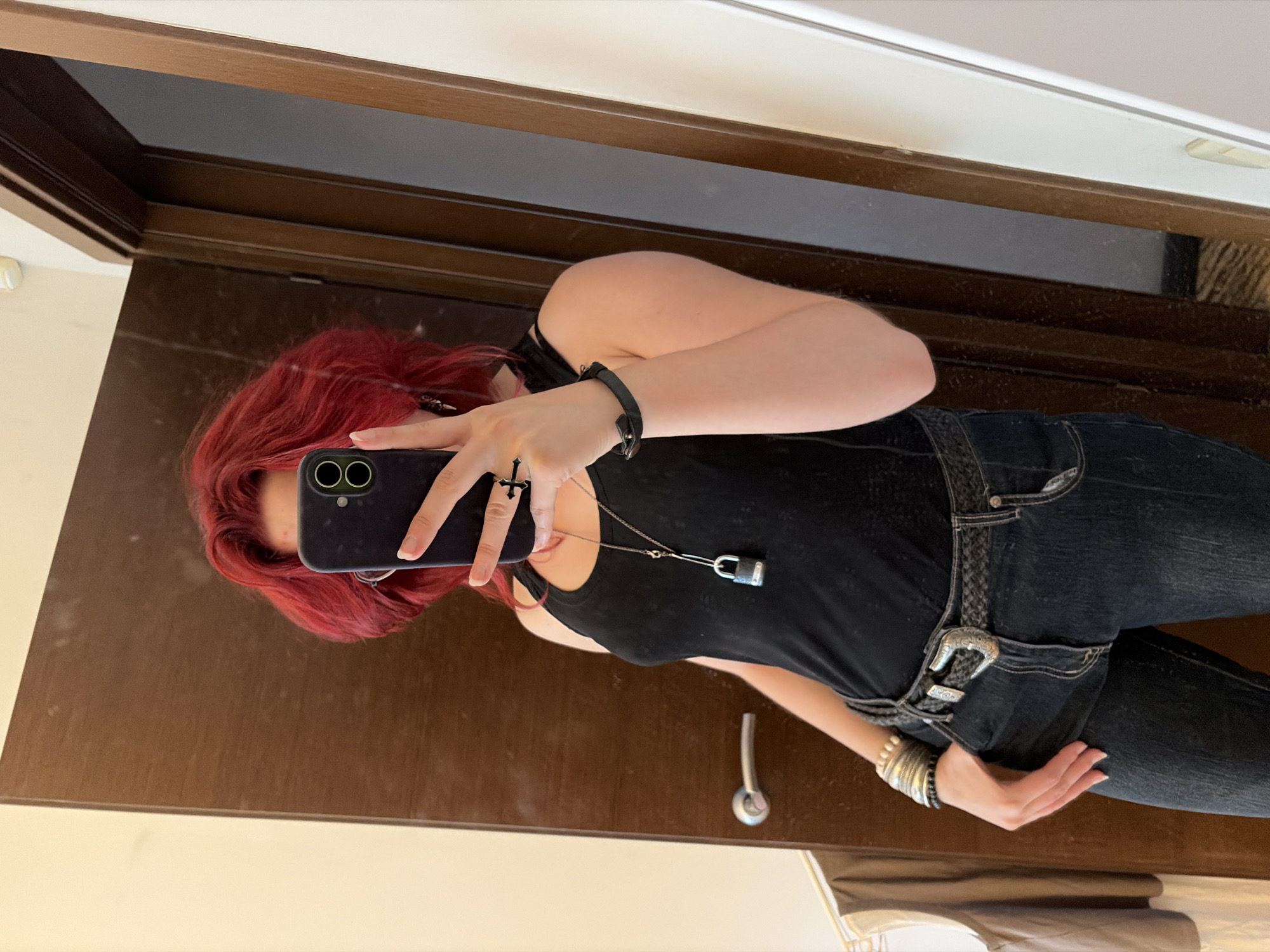 A person with bright red, voluminous hair is taking a mirror selfie in a room. They are wearing a black tank top, dark wash jeans, a braided belt with a large silver buckle, and multiple bracelets on their left wrist. A necklace with a silver padlock pendant is visible. The person is also wearing a ring with a cross design on their right hand. The background consists of a wooden door, a wall, and a glimpse of clothing hanging on a rack. The environment appears to be indoors, possibly a bedroom or dressing area. There is no visible indication of the location being in the city of Chelyabinsk, Russia.