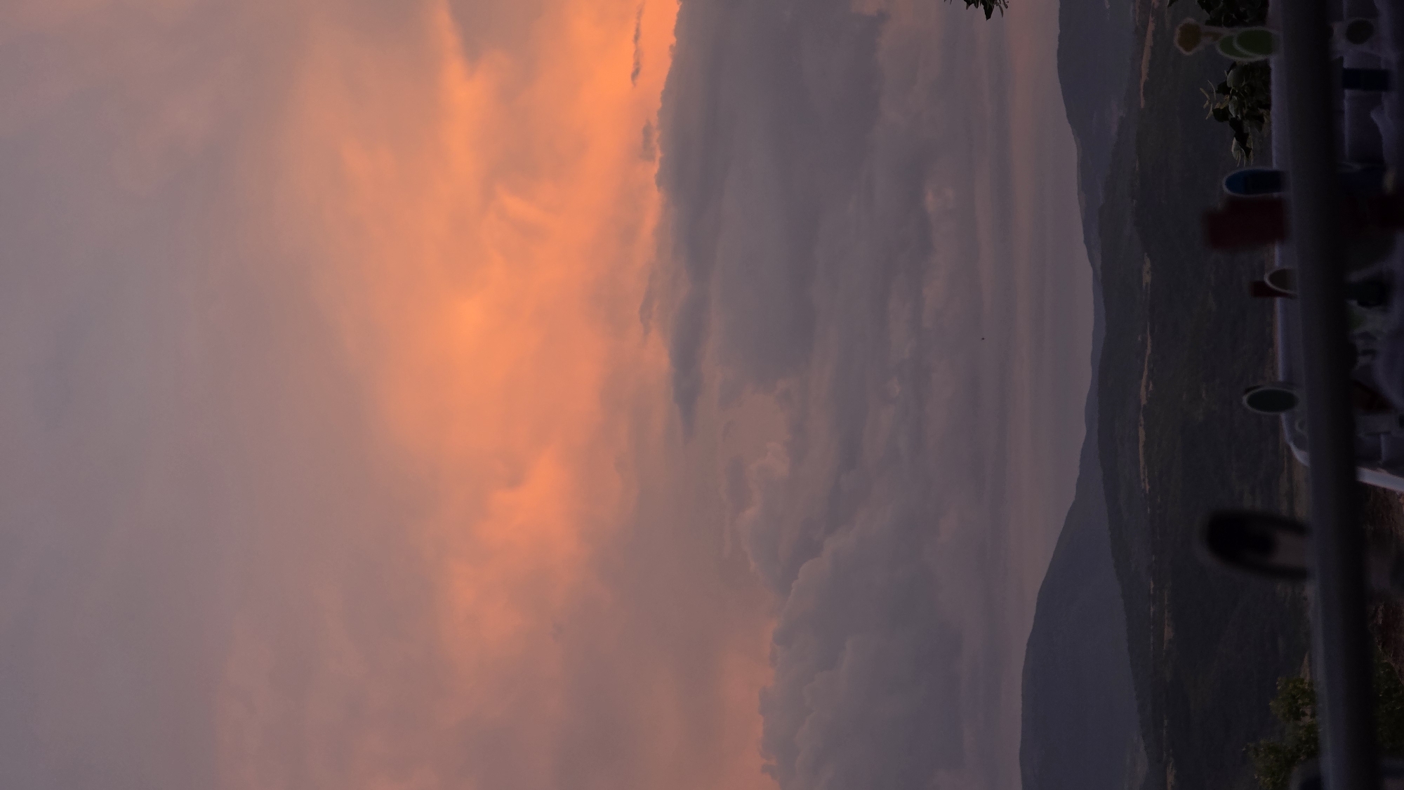 The image depicts a sunset over a mountainous landscape. The sky is filled with hues of orange and purple, with clouds scattered across the horizon. In the foreground, a railing is visible, with what appear to be clothespins or similar objects on it. The mountains in the background are dark and silhouetted against the colorful sky. The scene suggests a rural or semi-rural location, possibly a vantage point overlooking a valley. However, there is no specific visual information within the image that would allow for the identification of the city of Blagoevgrad, Bulgaria. The image focuses primarily on the natural elements of the landscape and the atmospheric conditions of the sunset. There are no visible people or specific actions being performed. The objects in the foreground are out of focus and do not provide detailed information.