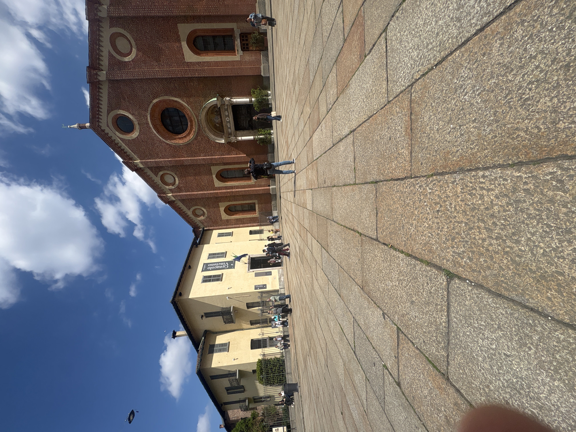 This image captures an outdoor scene in a public square, likely a piazza, bathed in bright daylight under a blue sky with scattered white clouds. The ground is paved with rectangular stone blocks in shades of light brown and grey.
On the right side of the frame, a grand red brick building dominates, featuring multiple round and arched windows with stone sills. Its facade displays intricate brickwork patterns and an ornate entrance. A small spire or statue is visible at the very top of this building, silhouetted against the sky.
On the left, a cream-colored building stands with several windows, some fitted with closed shutters, and a few small balconies. A large banner on this building prominently displays the text "CENACOLO VINCIANO", with "Milano" and "Santa Maria delle Grazie" also visible below it, indicating the location as the renowned site in Milan where Leonardo da Vinci's "The Last Supper" is housed.
Several people are scattered throughout the square. In the foreground, a man in a dark jacket and jeans stands, appearing to observe or photograph the architecture. Other individuals and small groups are visible further back, walking, standing, or congregating near the entrances of both buildings. A street light fixture is also visible against the sky.
The overall atmosphere suggests a casual public gathering, likely tourists or visitors exploring a historic and cultural landmark.