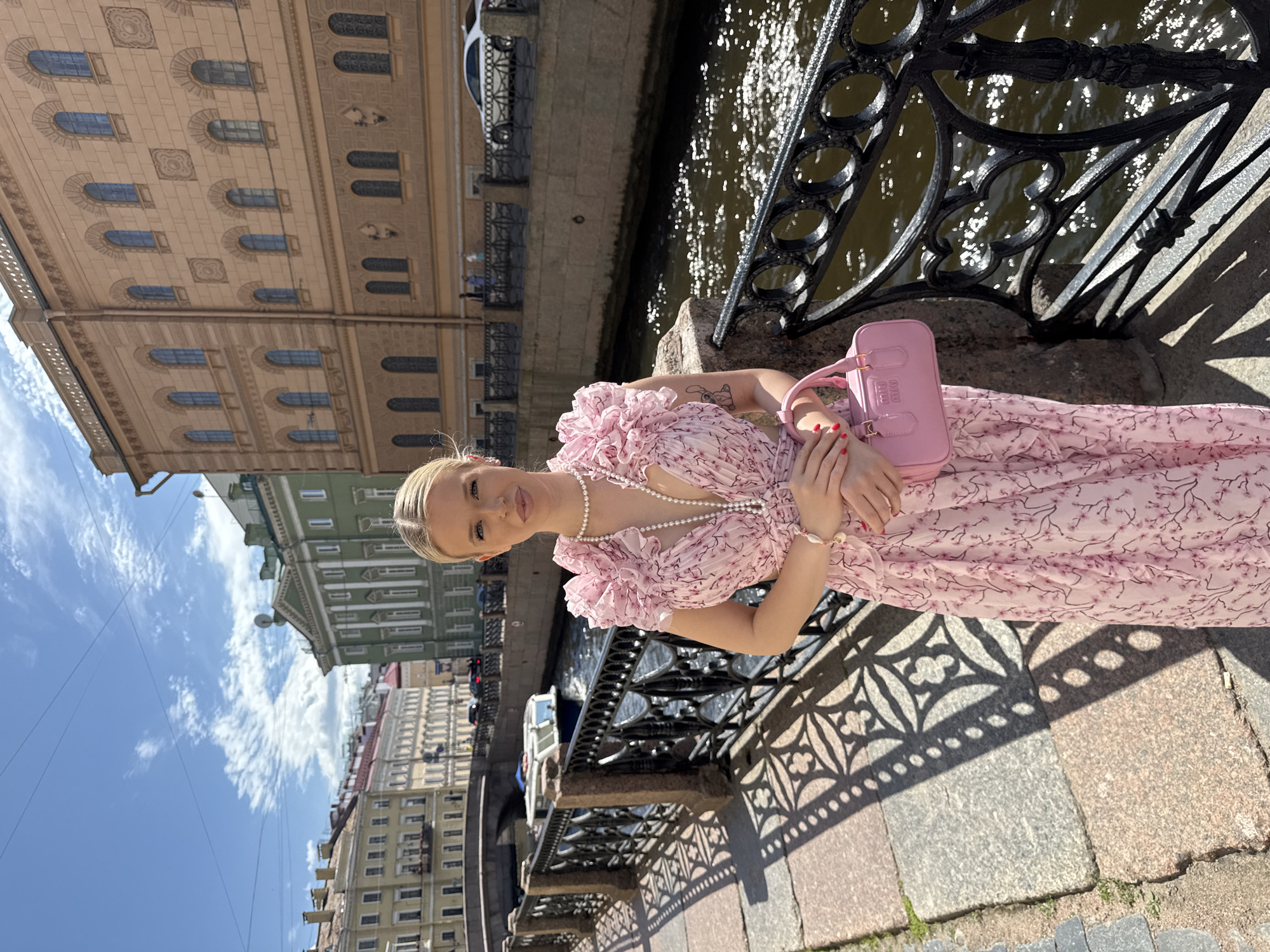 A young blonde woman in a light pink floral dress and a pearl necklace stands on a canal embankment in Saint Petersburg, Russia. She is holding a pink handbag. The woman is positioned on the left side of the frame, facing forward with a slight smile. Behind her, a dark canal reflects sunlight, and ornate black railings line the embankment. In the background, several historic buildings with classical architecture can be seen, with a bridge and boats visible on the canal. The sky is blue with scattered white clouds, and sunlight casts shadows from the railings onto the stone walkway. The overall scene depicts a sunny day in a picturesque urban environment.