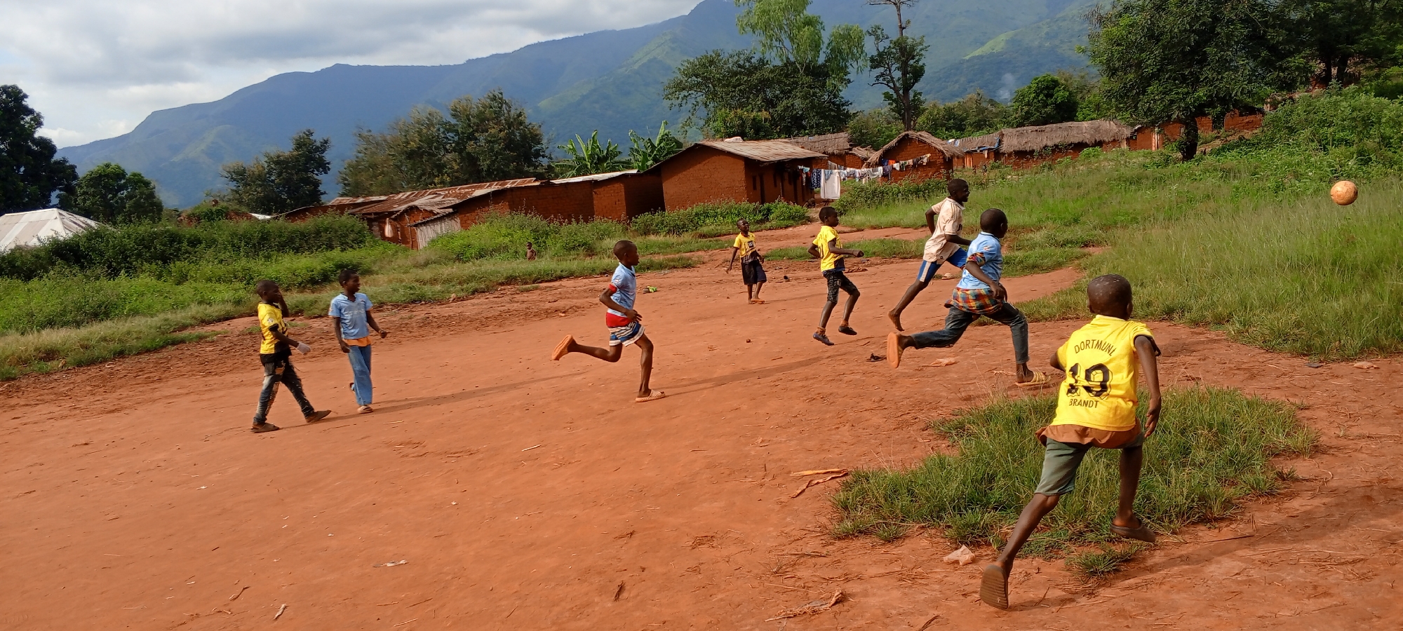 The image depicts a group of children playing soccer in a rural area of Ngalula, Democratic Republic of the Congo. The playing surface is an unpaved, reddish-brown dirt field. Seven children are visible, wearing a variety of clothing, including yellow and blue shirts. One child is wearing a yellow "Dortmund 19 Brandt" jersey. The children are in various stages of running and kicking. A soccer ball is in the air near the right side of the frame. In the background, simple brick or mud brick structures with corrugated metal roofs line the horizon. Laundry is hanging on a clothesline in front of one of the structures. Behind the village are rolling hills and mountains under a partly cloudy sky. Vegetation consists of grass, trees, and shrubs.