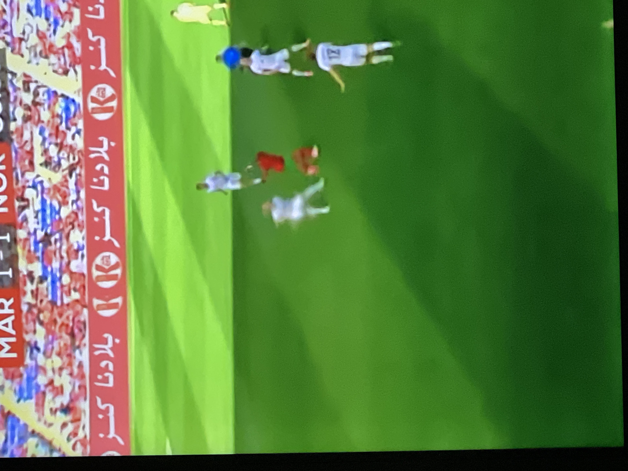 The image depicts an active soccer (football) match on a green grass pitch, likely held in a stadium in Casablanca, Morocco.
At the top, a partially visible scoreboard displays "MAR 1-1", indicating a score of 1-1, likely between Morocco and another team (the fragment "NOR" suggests Norway). Below the scoreboard, a red advertising banner spans the width of the field, featuring white Arabic text that reads "بلادنا كنز" (Biladuna Kanz), translating to "Our Country is a Treasure," alternating with a white "K" logo within a circle. Behind the banner, blurred figures of spectators in stadium seating are visible, predominantly in red, white, and blue hues.
On the vibrant green field, nine individuals are visible. Five players are in white uniforms (jerseys and shorts), with one player in the foreground wearing the number "12" on his back. Two players are in red uniforms (jerseys and shorts) and are currently on the ground, suggesting a fall or collision. A single player in a yellow/cream uniform, distinct from the white-clad players, is visible in the far upper-right, likely a goalkeeper. One individual wears a blue jersey and darker shorts, positioned near the fallen red-jersey players, indicating an official such as a referee.
The scene captures a moment of dynamic play or its immediate aftermath, with standing players observing the two fallen red-jersey players. The overall image quality is blurred and slightly grainy, consistent with a photograph taken of a screen displaying a fast-moving event. Dark vertical borders on the left and right indicate the original image source was a screen capture.