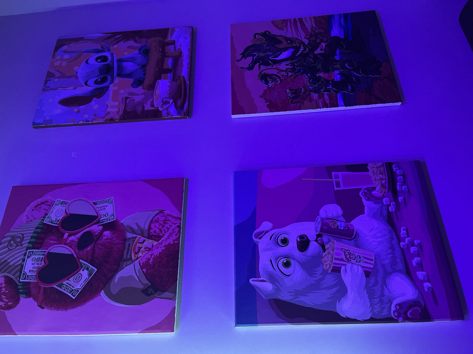 The media file captures four stylized paintings displayed on a wall in an indoor setting, likely in Kudrovo, Russia. The room is illuminated by a strong blue and purple light, possibly from LED strips, which bathes the wall and paintings in a distinct glow and altered color palette.
The top-left painting features Stitch from Disney's Lilo & Stitch, rendered in blue and purple hues, sitting beside a brown, wooden-like object and a white cup. The top-right painting depicts a dark, menacing creature resembling a dragon or monster with sharp teeth and claws, set against a background of deeper purples and reds.
The bottom-left painting shows a brown teddy bear wearing heart-shaped sunglasses and a green striped shirt, with light-colored shoes. Several one-hundred-dollar bills are draped over its sunglasses and body, with visible text including "FEDERAL RESERVE NOTE," "100," and "PULSMAS CURS." The bottom-right painting portrays a light-colored dog, possibly a bulldog or pug, lying on its back while holding a can with some text and a popcorn box that clearly displays "POP."
All four canvases appear to have edge lighting, enhancing the glow effect. The art style across the paintings is consistent, featuring bold outlines and distinct blocks of color. The ambient lighting creates a vibrant and somewhat surreal atmosphere.