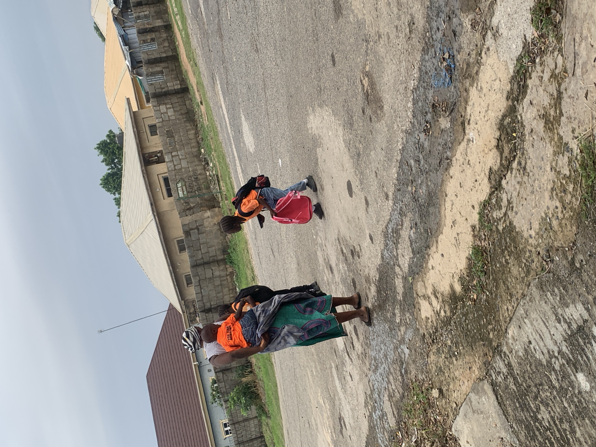 In Abuja, Nigeria, a woman in a headscarf and a patterned skirt carries a child on her back. She wears an orange shirt. To her right, a child in an orange shirt and blue jeans walks away, carrying a pink backpack and a small object in their hand. Further to the right, another child in an orange shirt and blue jeans walks towards the right, wearing a black backpack. They are on a paved road with some debris and puddles. In the background, buildings with various roof styles and a stone wall are visible. The sky is overcast.