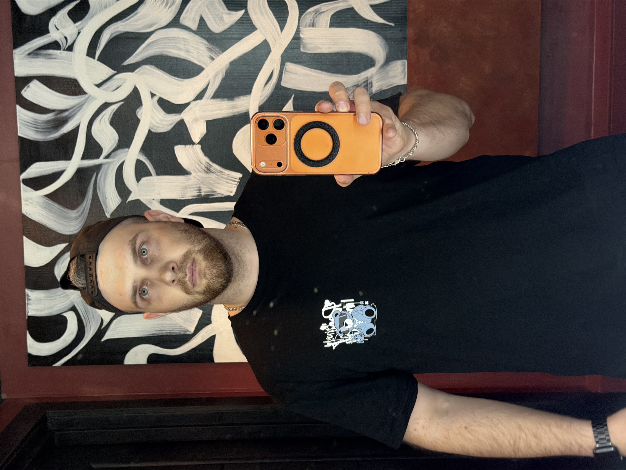 A man with light skin, light blue eyes, and a beard is taking a selfie in a dimly lit location. He is wearing a black t-shirt with a graphic on the left chest, a gold chain necklace, a brown baseball cap, and a silver watch on his left wrist. He is holding an orange iPhone with a black circle on the back in his right hand. Behind him is a black and white abstract painting in a red frame. Below him appears to be a dark wooden surface. There is no indication that this photo was taken in Izhevsk, Russia.