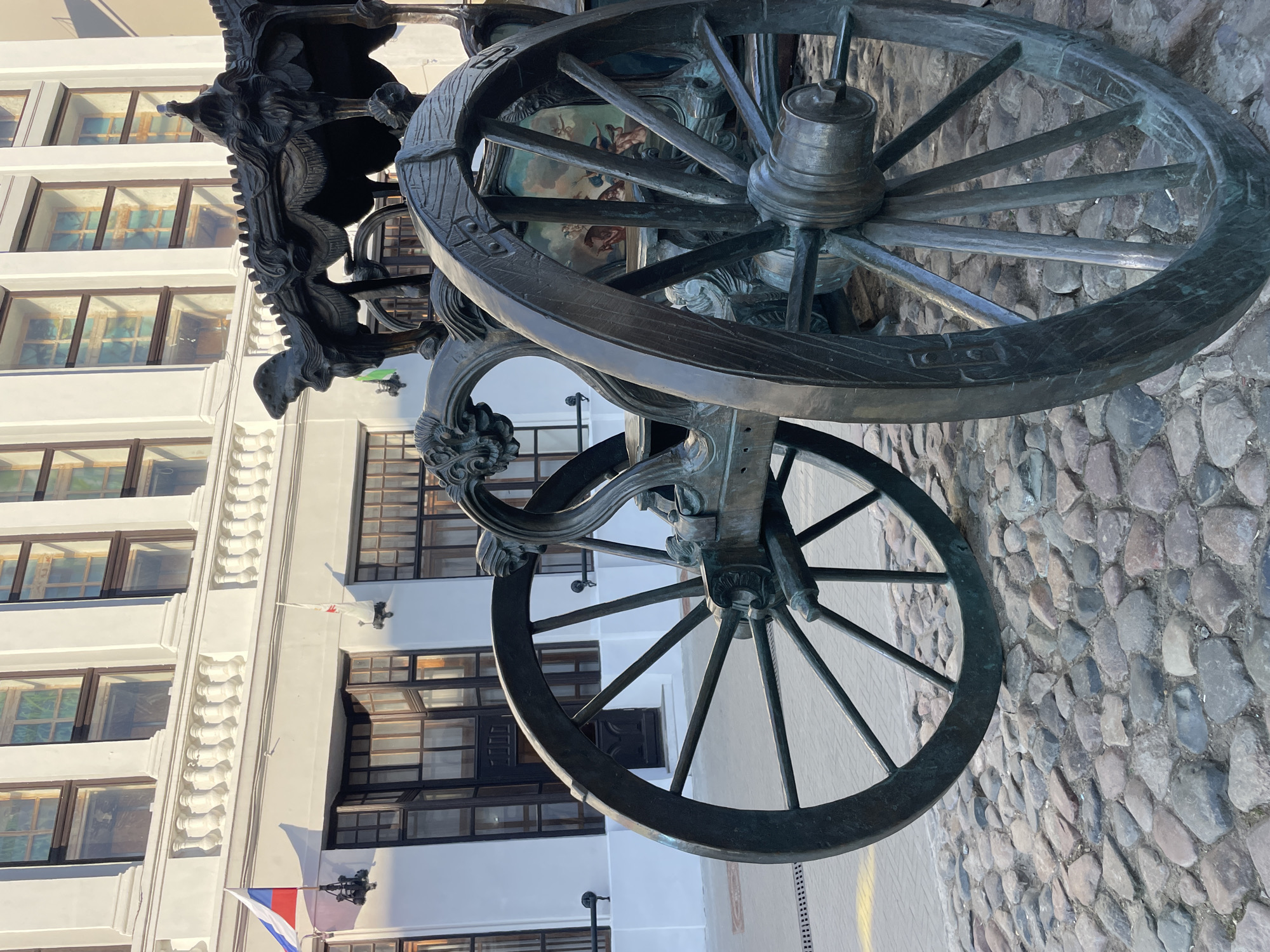 A detailed close-up shot features a bronze carriage standing on a cobblestone street, with a building in the backdrop. The carriage's wheels are intricately designed with spokes, and its frame boasts ornate carvings. Behind the carriage, a white building is visible, featuring multiple windows with dark frames. Two flags are attached to the building, one appearing to be the Russian flag. The photograph appears to have been taken in Kazan, Russia, focusing on the historical or artistic elements of the carriage against the backdrop of the city's architecture.