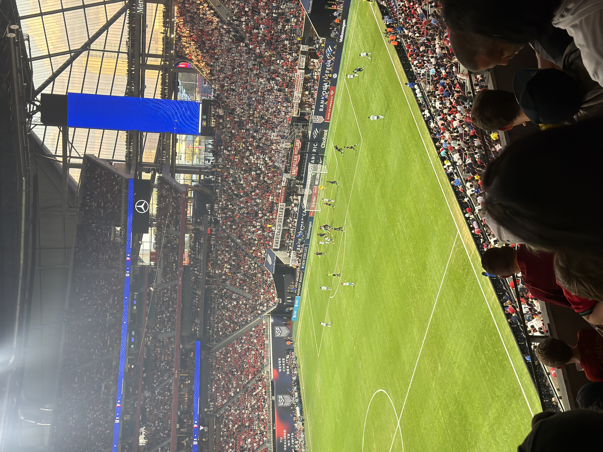 This image depicts a soccer game taking place in a stadium in Atlanta, United States of America. The stadium is filled with spectators, predominantly wearing red and white, watching the game. A large blue video screen is visible above the field, as well as stadium advertisement branding. People are visible in the lower right foreground, likely spectators at the game. The environment suggests a well-attended sporting event.