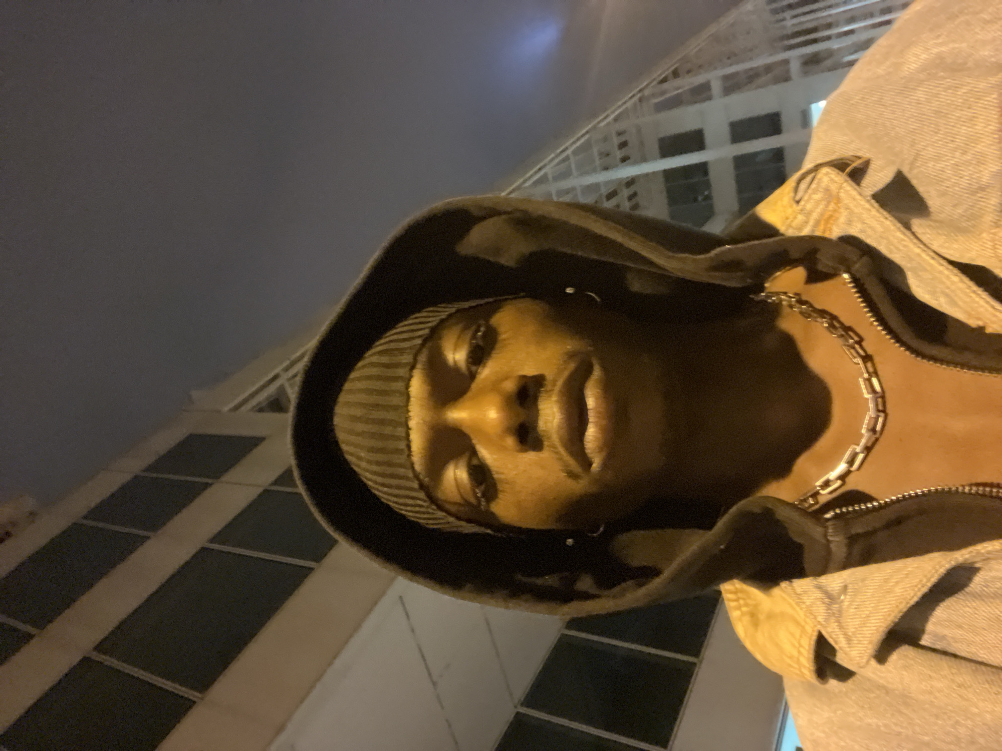 A medium shot looking up at a person wearing a knit cap and a hooded jacket, against a backdrop of tall buildings. The person is positioned in the lower center of the frame, with their face turned towards the viewer. The hood is up and frames their face. The person is also wearing a chain necklace. A section of a light-colored jacket is visible on the right side of the frame. In the background are tall buildings with dark windows. The sky is dark and overcast. The city is not identifiable as Barcelona.