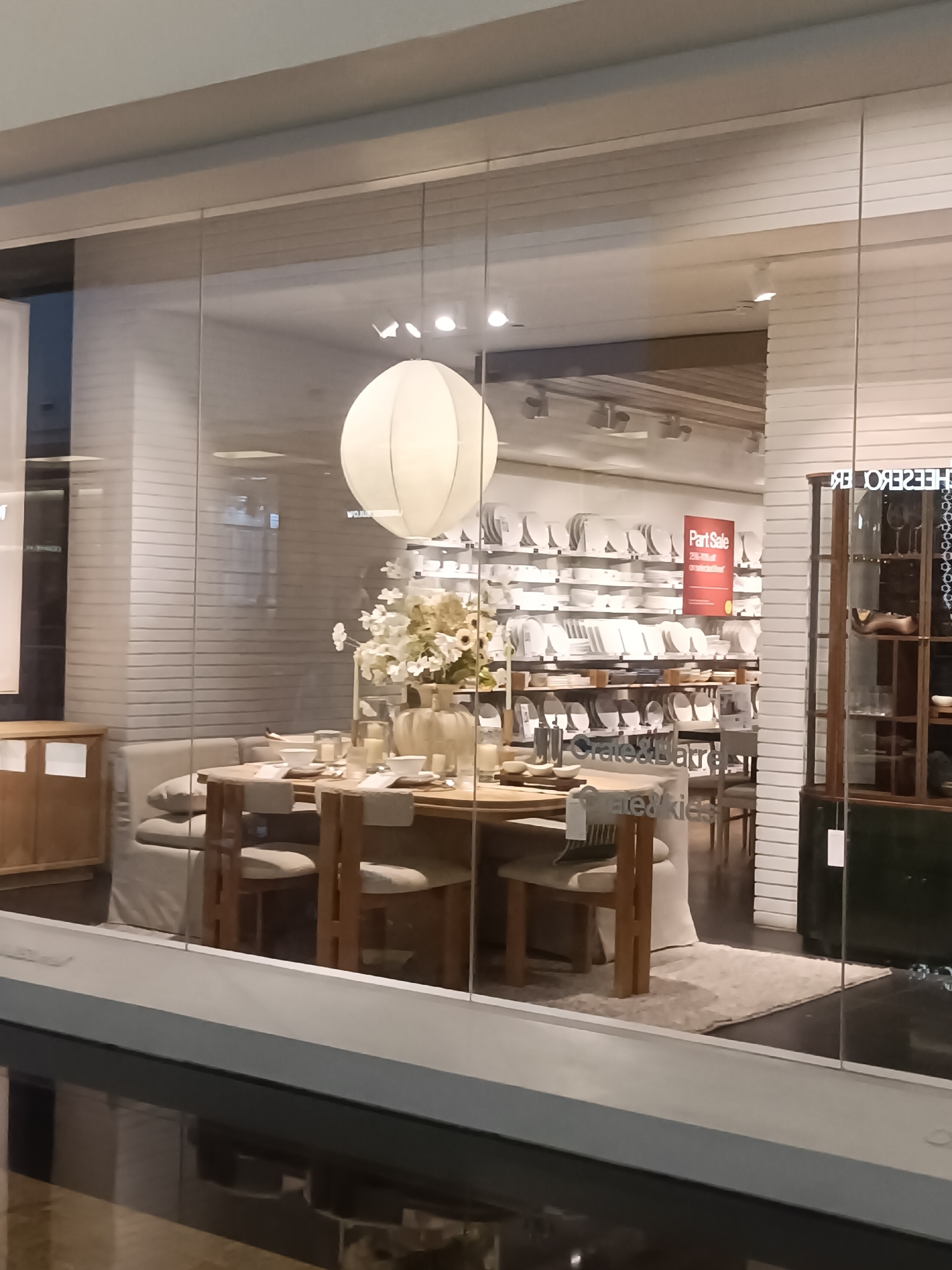 A storefront display for Crate & Barrel, a home furnishings retailer, is visible through large glass windows. The display features a dining table set for a meal, with chairs and place settings. Behind the table, shelves are stocked with white plates and bowls. A large, white, globe-shaped pendant light hangs from the ceiling. A red "Part Sale" sign is prominently displayed. The store appears to be located in a mall, with reflections of other stores visible on the glass. The scene is clean and well-lit, suggesting a retail environment. There are no people visible in the display. The overall impression is one of an upscale, modern home goods store.