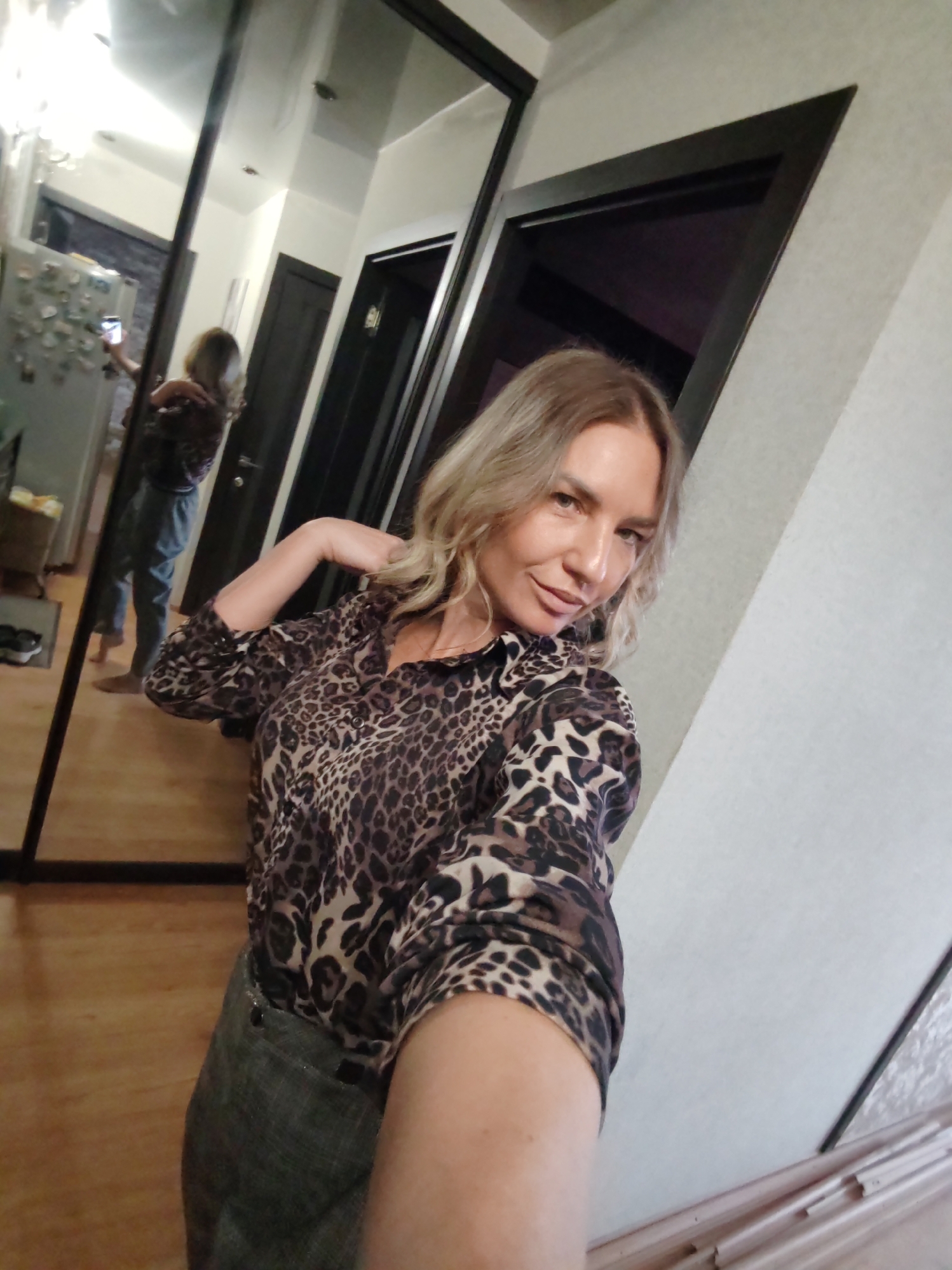 The image captures a woman taking a mirror selfie indoors, likely in a residence in Petropavlovsk-Kamchatsky, Russia. The woman, with blonde, wavy, medium-length hair, is seen from the chest up in the foreground. She wears a long-sleeved leopard print blouse in shades of brown and black. Her right arm is bent, with her hand near her neck, and she looks directly at the camera with a slight smile.
Her reflection is visible in a large, dark-framed mirror behind her, showing her full body. In the reflection, she is holding a smartphone up with her left hand to take the photo. She is wearing light blue jeans and appears to be barefoot.
The background of the reflection reveals a hallway with a light wooden floor, a white refrigerator covered with various magnets, and a light-colored wall. Two dark-colored doors are also visible in the background, one in the main view and one in the reflection, suggesting an interior hallway or entryway. The setting is illuminated by indoor artificial lighting. No text is visible in the image.