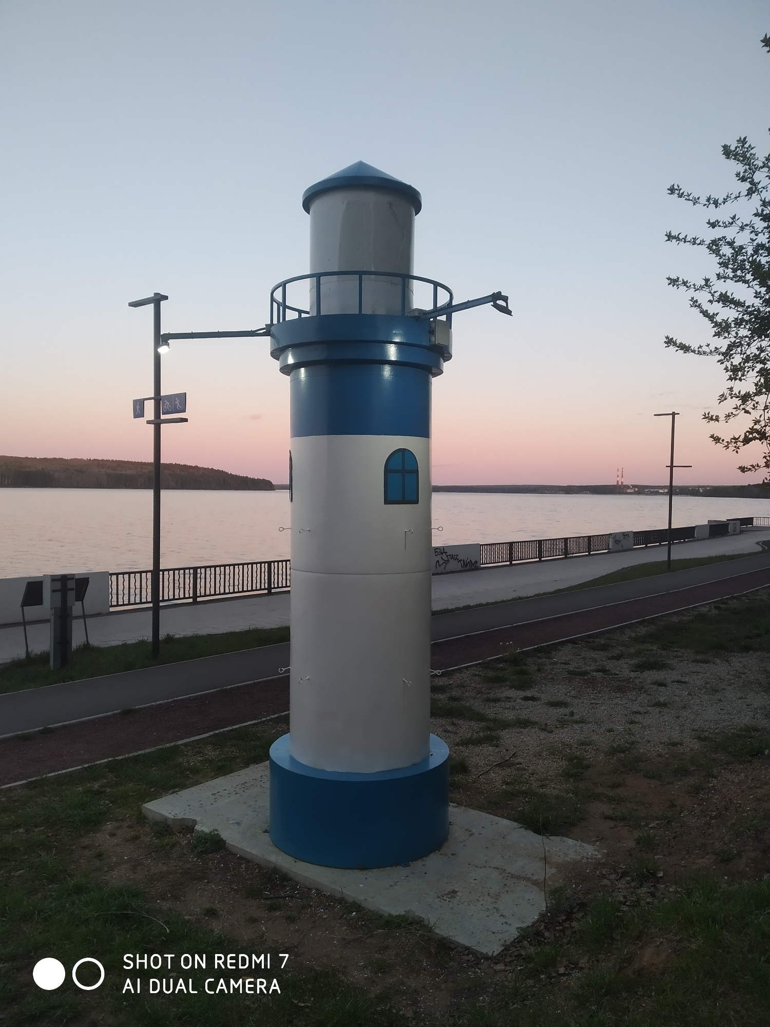 The image shows a decorative lighthouse located on a promenade in the city of Chaikovsky, Russia. The lighthouse is painted white and blue, featuring a cylindrical tower with a round blue base and top, a white middle section with a small arched window, and a top section with a railing. The lighthouse sits on a concrete base amidst a grassy area. Behind the lighthouse, there is a waterfront view with a body of water, a low-lying treed shoreline in the distance, and utility poles and lampposts. There is a pedestrian path running alongside the water, with a black metal railing separating the path from the water. The sky is a gradient of colors ranging from blue to pink. A street sign is visible in the upper left, and a tree with green leaves is in the upper right. The bottom of the image has a camera watermark.