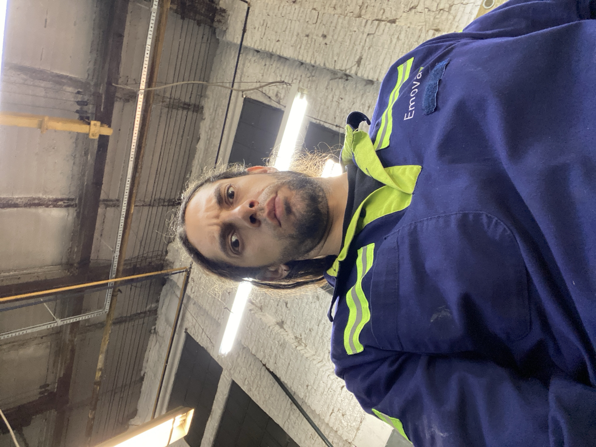 A low-angle, forensic portrait captures a male individual with long dark hair tied back and a beard, looking directly into the camera with a direct, unsmiling expression. He is wearing a dark blue work uniform featuring a lime green collar and lime green reflective stripes across the shoulders. The word "Emova" is embroidered in white on the right chest area of the uniform, above a dark blue Velcro patch. The uniform exhibits minor wear and light smudges. The background consists of an indoor industrial or workshop setting with rough, white-painted brick walls. The ceiling is composed of concrete and exposed metal beams, along with visible utility infrastructure including yellow and blue pipes and electrical conduits. Bright fluorescent tube lights are mounted overhead, illuminating the scene. Based on the "Emova" branding, the location is an operational or maintenance facility belonging to Emova, the company responsible for the Buenos Aires Underground (Subte) system, situated within Buenos Aires, Argentina.