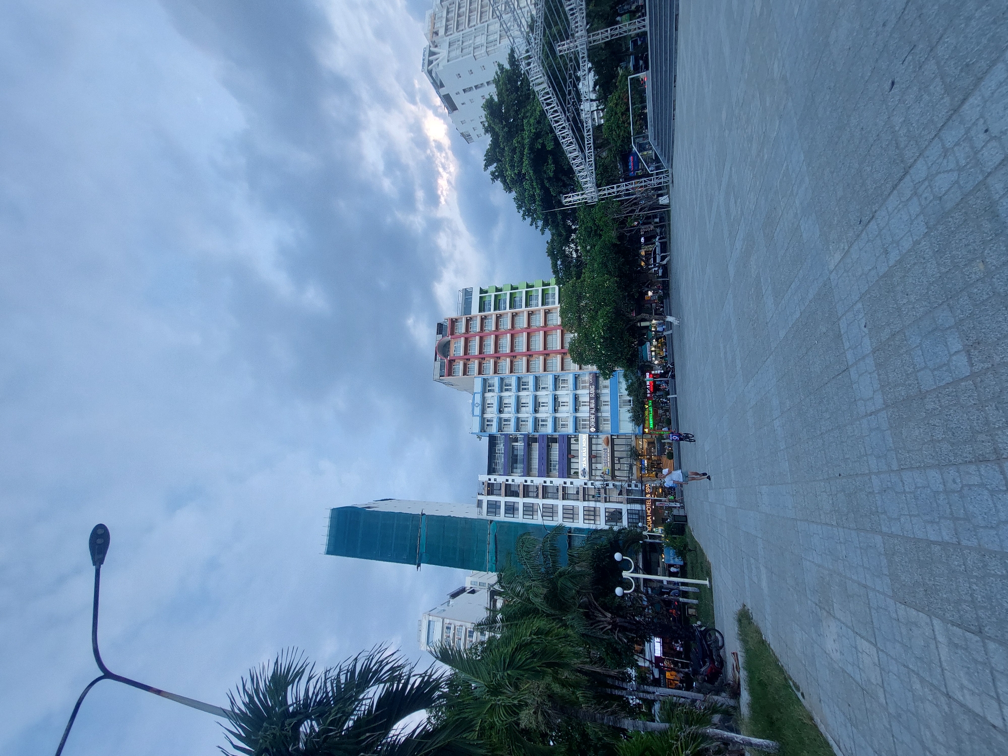 In Nha Trang Ward, Vietnam, this image captures an outdoor urban scene under a cloudy sky. A wide, paved plaza occupies the foreground, leading towards a row of multi-story buildings. A lone person is walking across the plaza, and another individual is riding a bicycle. To the left, palm trees and a street lamp line the edge of the plaza. The buildings display a variety of architectural styles and colors, with some undergoing construction, indicated by scaffolding. A large, metallic structure, possibly a stage or canopy, is visible on the right side of the frame, with steps leading up to it. The overall atmosphere suggests an evening setting, with the sky overcast and the lighting subdued.