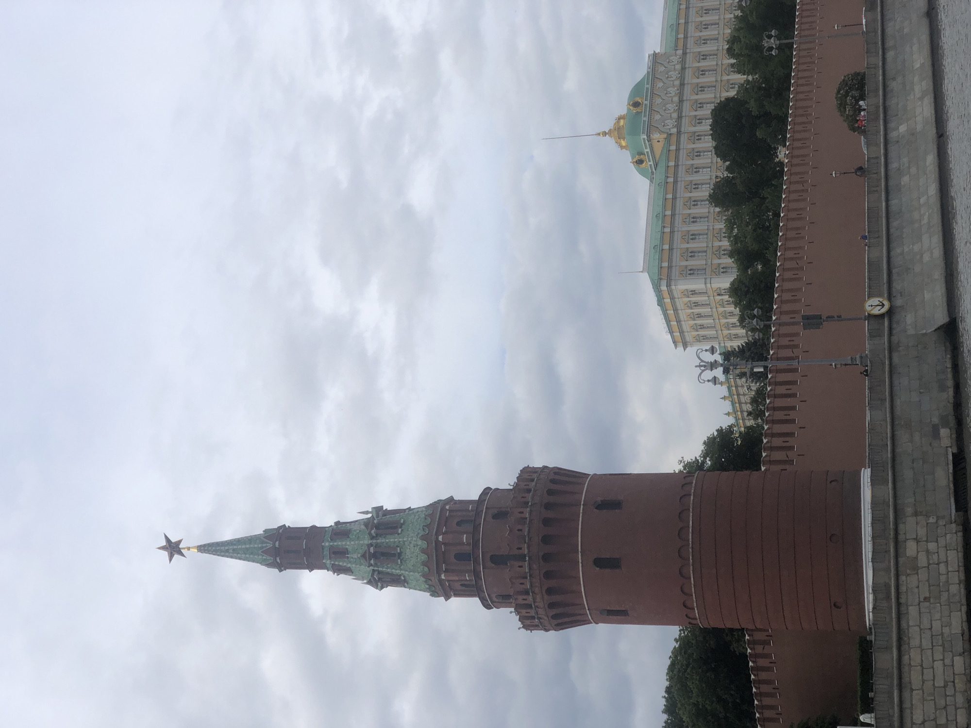 The image shows a view of the Moscow Kremlin, specifically the Kremlin wall and one of its towers topped with a red star. Part of the Grand Kremlin Palace with its golden domes is also visible. The weather appears overcast, with a cloudy sky. The perspective is from a lower angle, looking upwards at the structures. The location is Moscow, Russia.