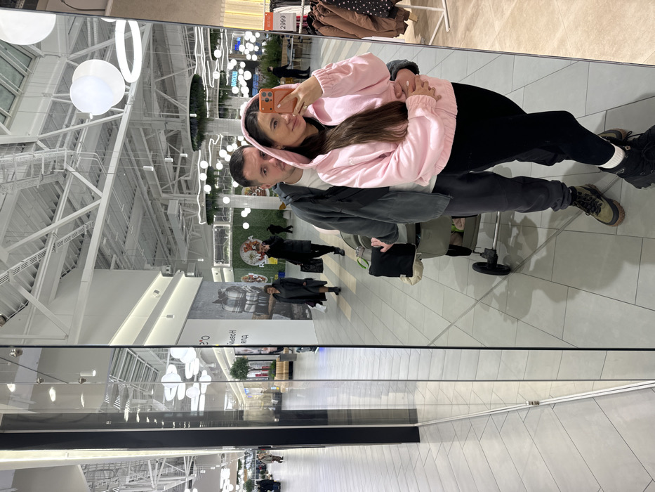 The image captures a selfie reflection of a couple and a baby stroller within a modern shopping mall, reportedly located in Yudino, Russia.
In the foreground, a woman, facing the camera's reflection, smiles while holding an orange-cased smartphone in her right hand to take the photo. She wears a pink hooded jacket over a black top, black leggings, and black boots. Her long dark hair is down. A man stands behind her, his right arm around her waist. He wears a dark grey hooded jacket, dark grey sweatpants, and olive green hiking-style boots, looking forward past the camera. A beige and black baby stroller is positioned to the left of the man, partially visible.
The environment is a brightly lit, spacious shopping mall interior with a high white ceiling featuring exposed structural elements, ductwork, and various suspended lighting fixtures, including white spherical and ring-shaped lights. The floor consists of light-toned geometric tiles. To the left, a series of reflective panels and pillars mirror parts of the mall. In the mid-ground background, reflected and direct views show multiple shoppers walking. A large banner with Russian text, including "Новый год" (New Year), is visible on a store facade. To the far right, a clothing rack displays apparel with a red "2999" price tag. Other retail displays and decorative green elements are scattered throughout the background.