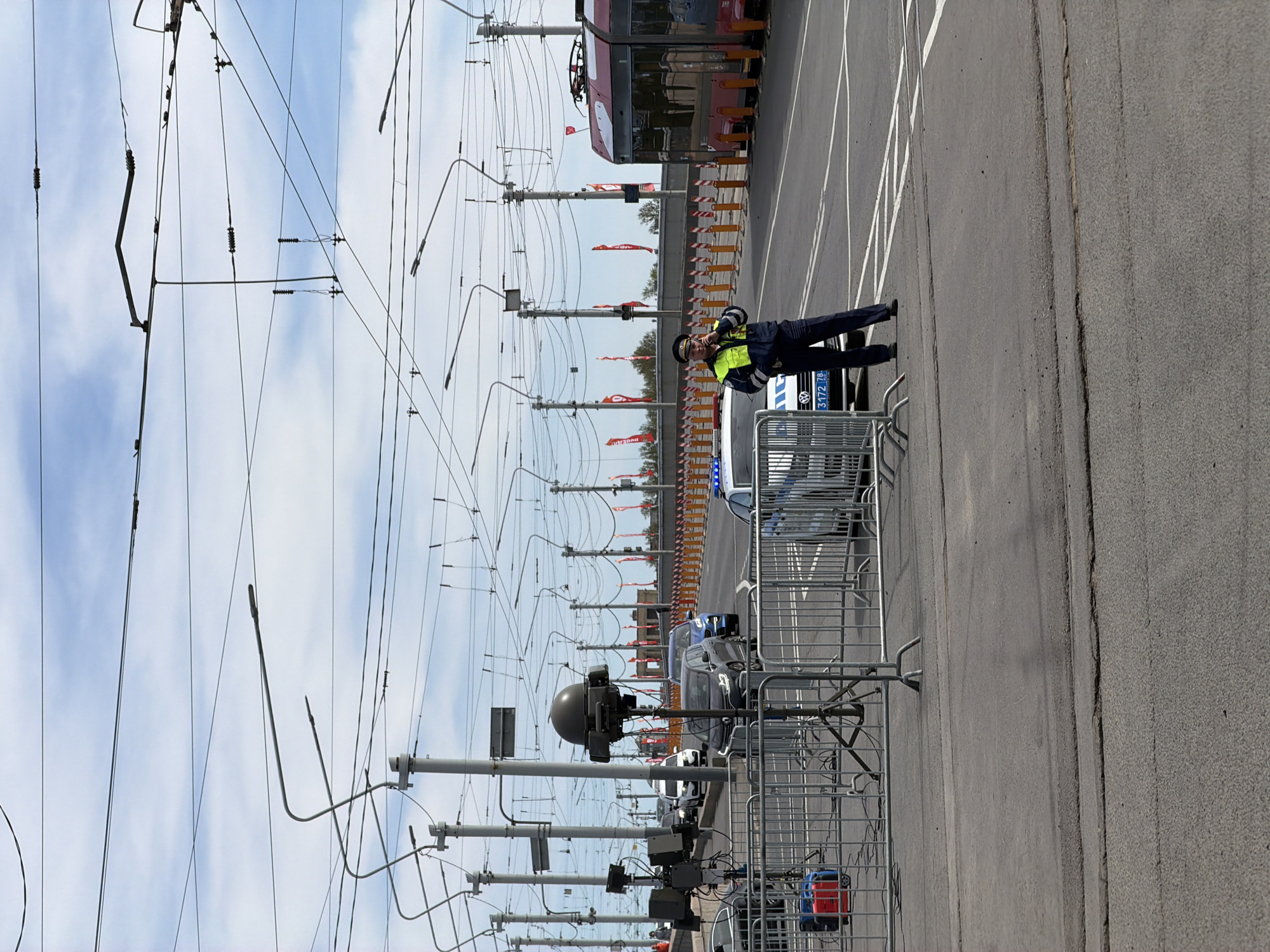 The image depicts a street scene in Saint Petersburg, Russia. A police officer, dressed in a dark blue uniform with a yellow high-visibility vest, is standing next to a silver and blue police car. The vehicle is behind a metal barricade. Overhead are numerous electrical lines and poles, typical for trolleybus or tram systems. Red and white flags are strung along the poles in the background. A red trolleybus is also visible in the background. The street is paved with asphalt, marked with white lines. The sky is blue with scattered white clouds.