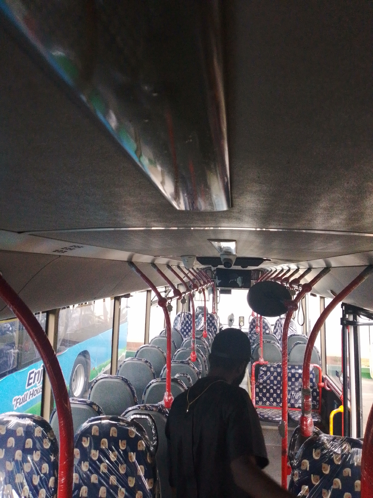 The image captures the interior of a bus, viewed from a low angle looking towards the front. The ceiling of the bus is visible, with a reflective strip running along its length. Below the ceiling, a series of red poles and horizontal bars are present, forming a railing system for passengers. The seats are mostly covered in clear plastic, with some featuring a blue fabric with a white circular pattern. A man wearing a dark t-shirt and a cap is standing in the aisle, facing away from the camera. He appears to be walking towards the front of the bus. Through the windows on the side of the bus, a blue exterior with white text that appears to read "Enjoy Full Hous" can be seen. The overall lighting suggests it is daytime, with light entering from the windows. The location is identified as Barikin Jirga Summa, Nigeria.