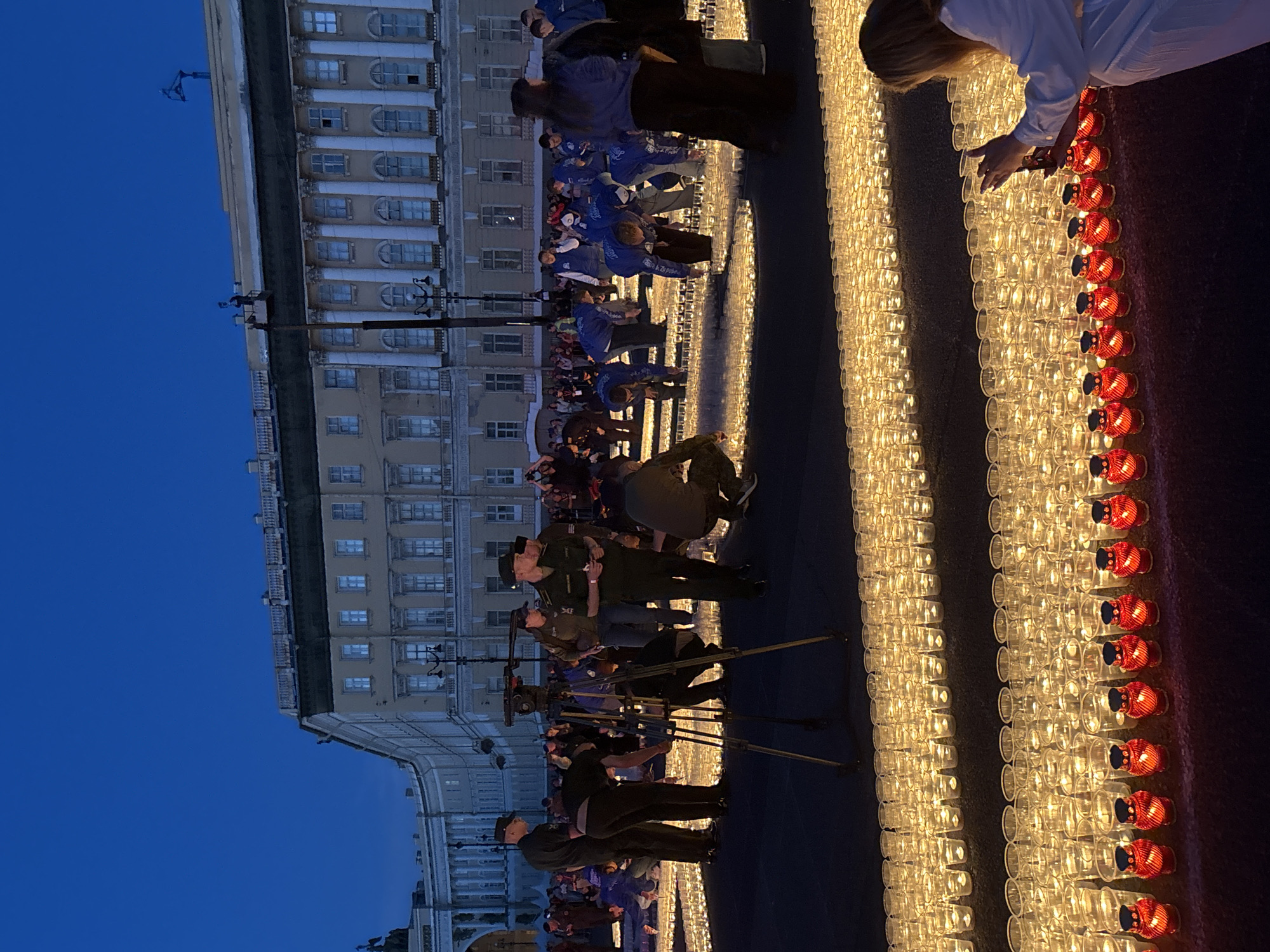 The image captures a somber event taking place at dusk in Palace Square, Saint Petersburg, Russia. A vast field of illuminated votive candles stretches across the foreground, arranged in neat rows. Many people, some in uniform and others in casual attire, are seen bending down and tending to the candles, suggesting they are being lit or arranged. The imposing classical architecture of the General Staff Building, a landmark of Palace Square, forms the backdrop, its facade illuminated by the fading daylight. The sky above is a deep blue, indicating the transition from day to night. The scene is peaceful yet solemn, with the glowing candles creating a profound atmosphere.