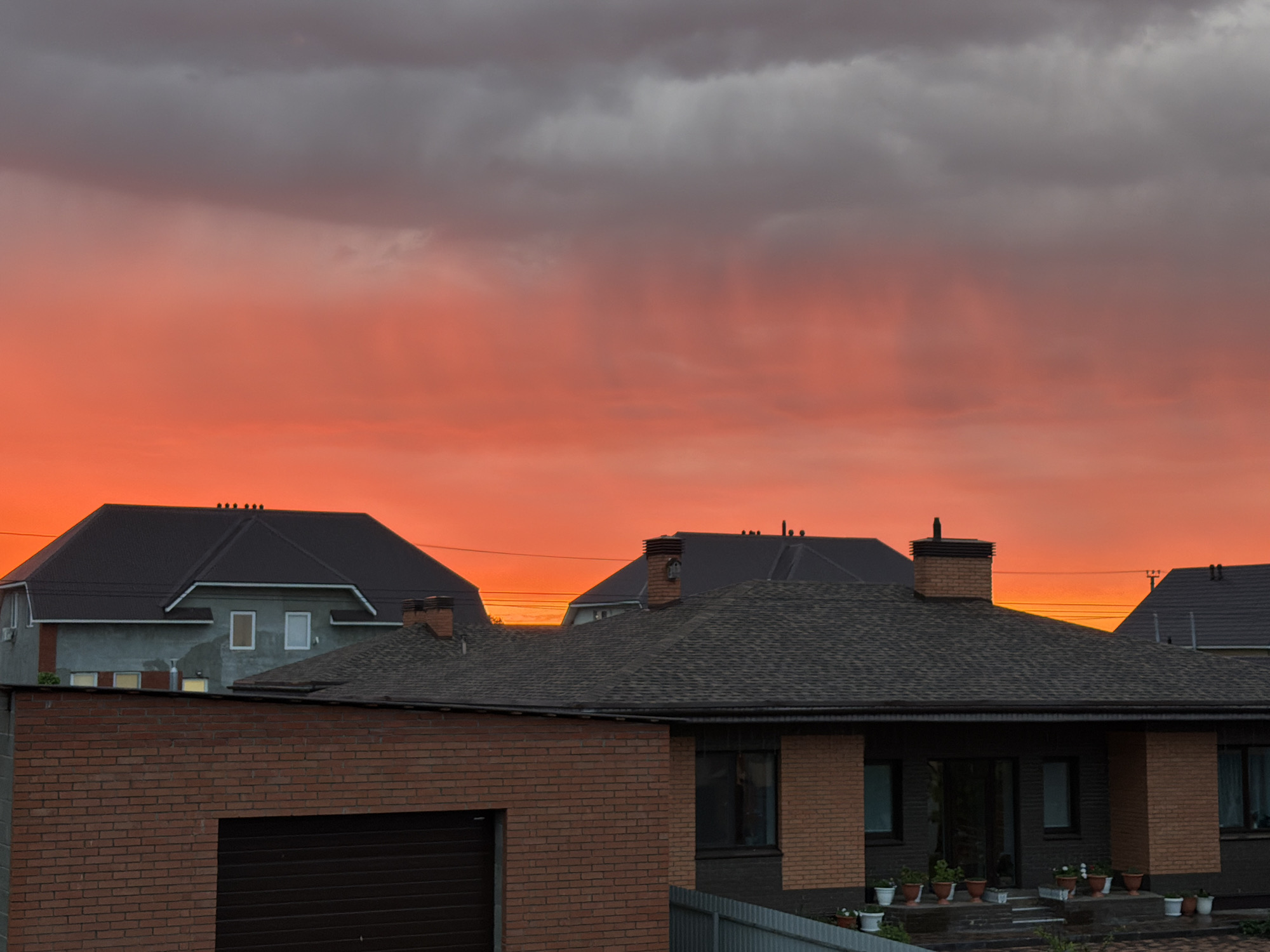 The image shows a residential area in Большая Шильна, Russia, at dusk or dawn, as evidenced by the vibrant orange and gray sky. Several houses are visible, featuring dark, gabled roofs and brick or gray-painted walls. One house has a brick garage and a variety of potted plants on its porch. An adjacent house shows its gray walls and dark roof as it's situated directly behind the brick house with potted plants. Power lines are strung across the sky between the rooftops. A fence runs across the lower portion of the image.