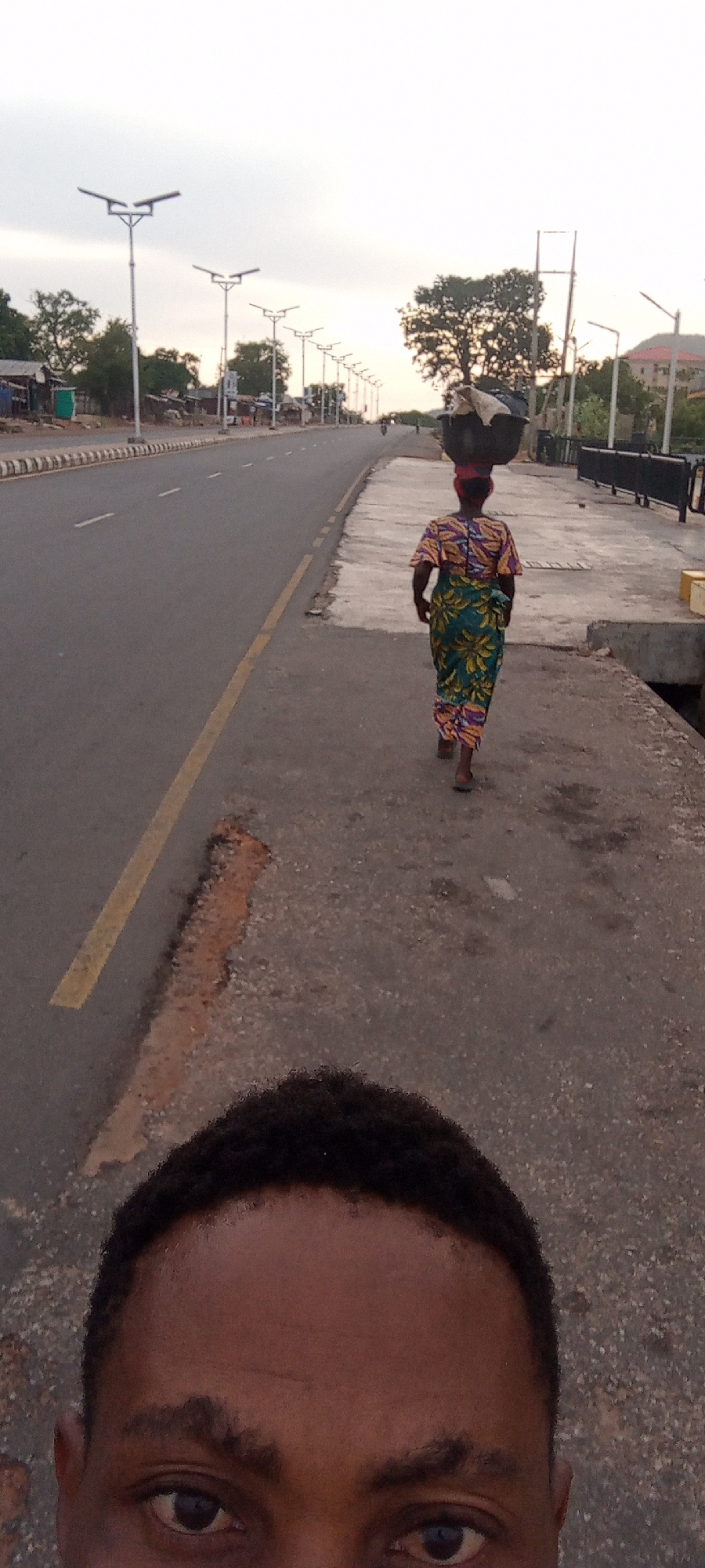 A street-level photograph taken in Adankolo, Nigeria, shows a partially visible person in the foreground. Behind them, a woman walks along a concrete sidewalk, carrying a container on her head. A paved road with street lights runs parallel to the sidewalk. Trees and buildings can be seen in the background.