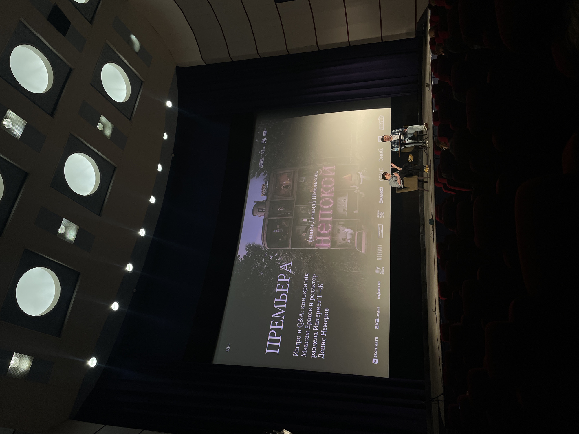 The image shows a cinema screen with a film title overlayed on a dark background, possibly during a premiere or a Q&A session. Two men are seated on chairs on a stage in front of the screen, facing the audience. The man on the left is wearing a grey shirt and dark pants, gesturing with his hands. The man on the right is wearing a checkered shirt and blue jeans, looking towards the man on the left. The cinema hall appears to be dimly lit, with rows of red seats visible in the foreground, mostly in shadow. The screen displays text in Russian, including "ПРЕМЬЕРА" (Premiere) and what appears to be film credits and logos of various media outlets at the bottom. The ceiling of the cinema features an unusual design with circular and rectangular light fixtures. There is no specific information in the image that identifies the location within Saint Petersburg, Russia.