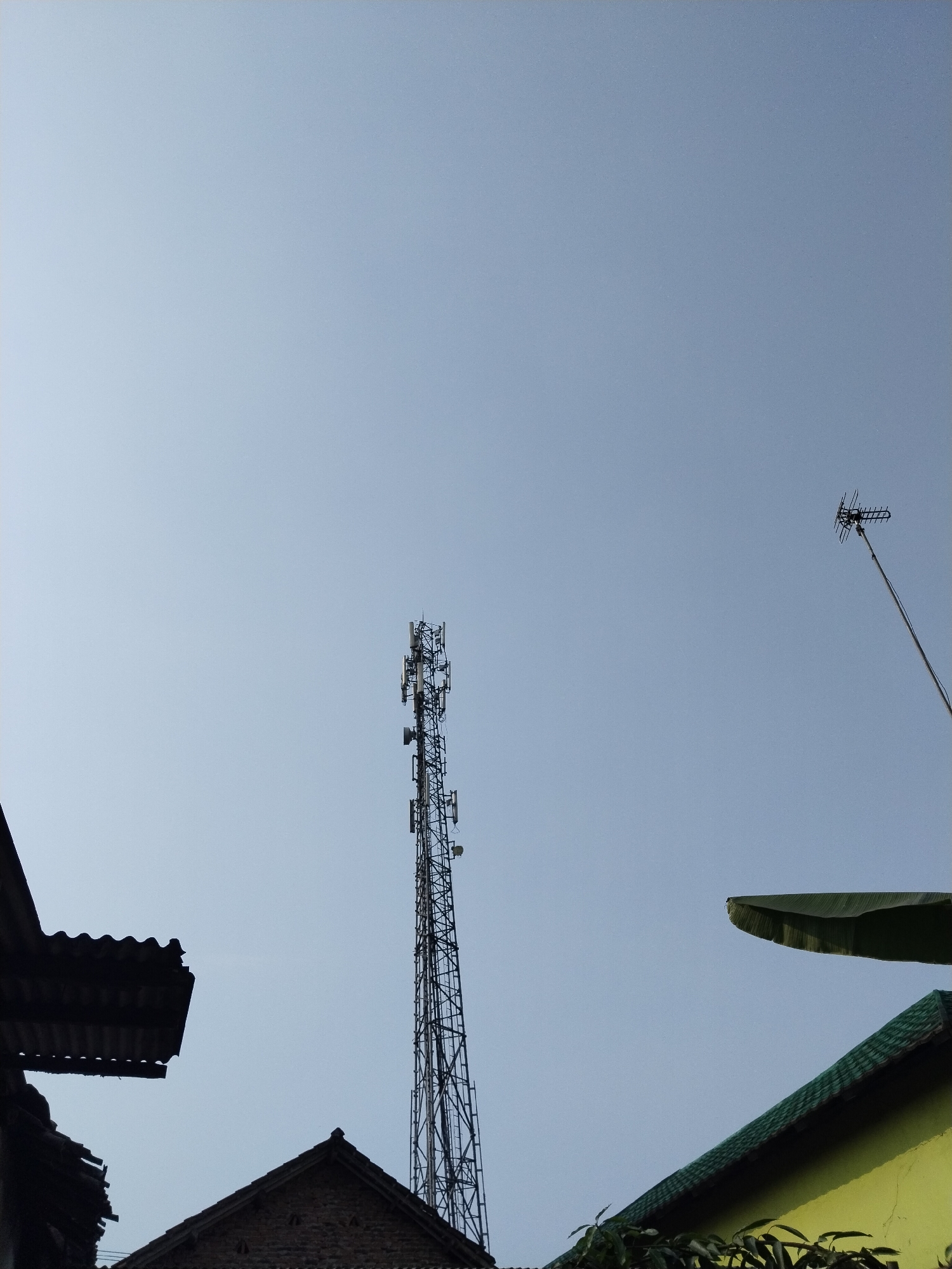 This is a low-angle, outdoor, daytime shot of a cell tower and a television antenna silhouetted against a clear, pale blue sky. The image appears to be taken in Kampung Baru, Indonesia, given the context provided. In the foreground, the dark silhouettes of rooftops are visible on the left and right sides of the frame, with a yellow building and green roof also present on the right. The cell tower is centrally located and is a tall, lattice-like metal structure with multiple antennas attached. To the right, a television antenna extends from a thin pole. There are no people or animals visible in the scene. The overall impression is one of urban infrastructure against a serene sky.
