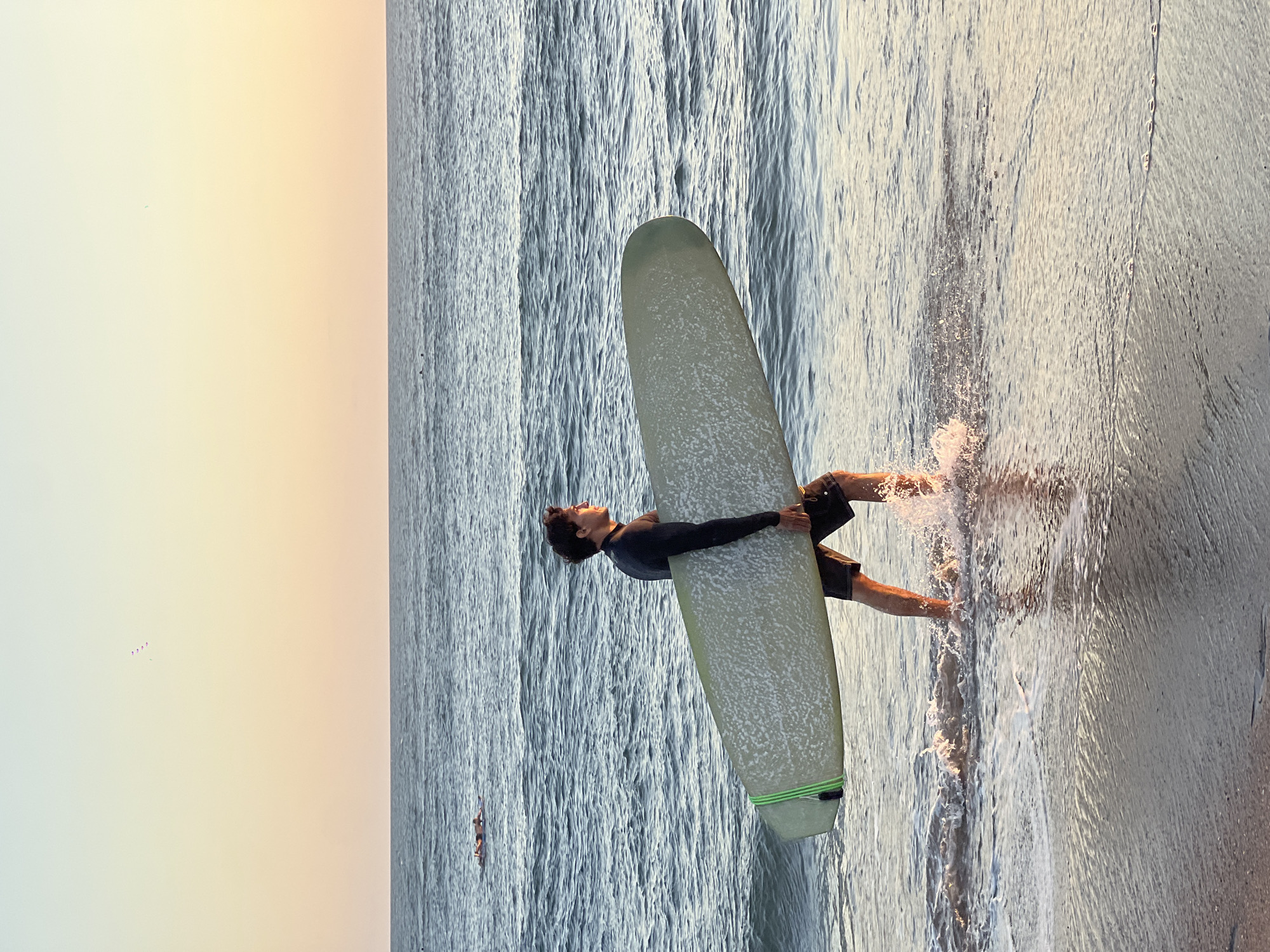 The image depicts a person standing in the water near the shoreline, holding a surfboard. The person is wearing a dark-colored long-sleeved shirt and dark shorts. The surfboard appears light-colored with a green accent on the tail. Splashes of water surround the person's legs and feet, indicating movement or shallow water. The background shows the ocean with visible wave patterns. In the distance, there is another person or object visible in the water. The sky is bright, with a gradient of light colors suggesting sunrise or sunset. The location is likely North Kuta, Indonesia, given the tropical coastal environment and common surfing activity in that region.