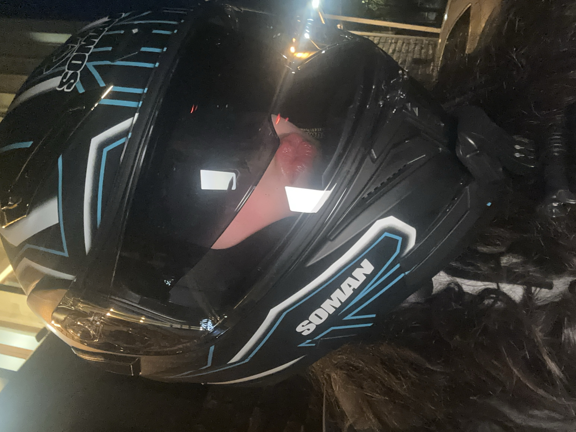 A person's head, primarily their dark, curly hair, is visible at the bottom right of the frame, with a black motorcycle helmet resting on or being worn by them. The helmet is the central subject, prominently featuring a sleek, angular design with white and light blue graphic accents on its black shell. The brand name "SOMAN" is clearly visible in white uppercase letters with blue outlining on the side of the helmet.
The clear visor of the helmet reflects several bright artificial light sources, indicating that the image was taken at night or in a low-light environment. These strong reflections obscure any view of the wearer's face inside the helmet. The background is dark and largely out of focus, with some indistinct shapes and lights suggesting an outdoor setting, possibly urban, in Yerevan, Armenia. The overall impression is a nocturnal shot, highlighting the helmet's design and reflecting lights.