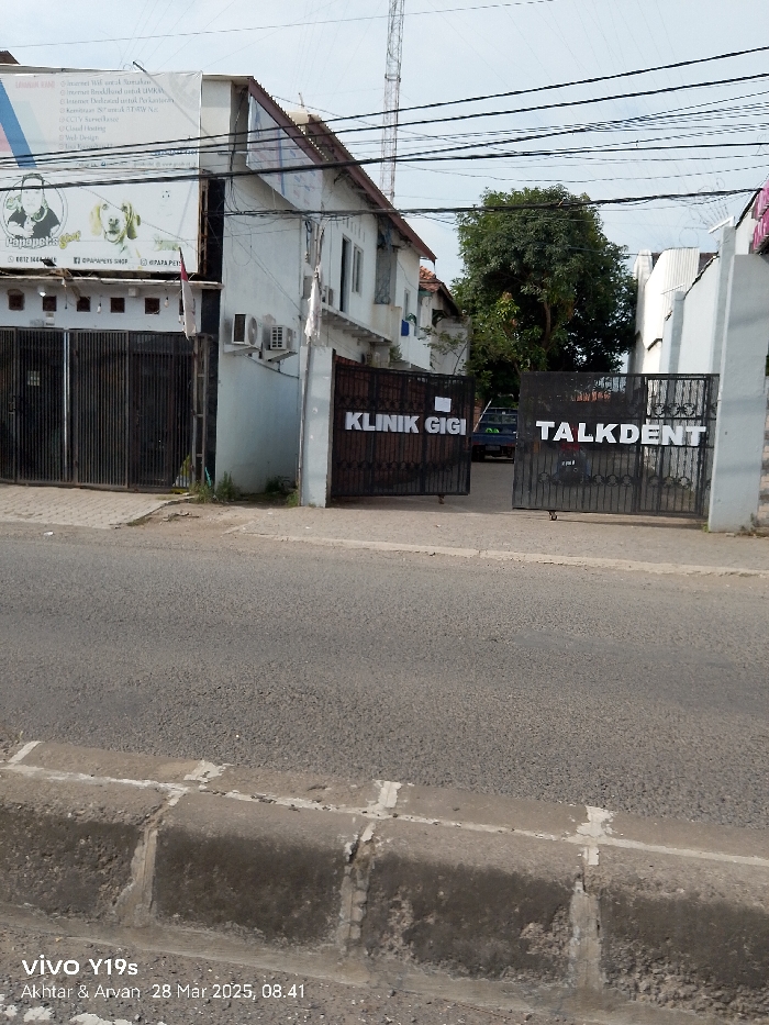 The image shows a street in Jatibarang, Indonesia. It is a paved road with a sidewalk on the left side. The street is lined with buildings, one of which is a dental clinic. The clinic has a metal gate with the sign "Klinik Gigi" written on it. Next to the clinic gate, there is a second gate with "Talkdent" written on it. A vehicle is partially visible in the background, parked inside the gated area. The sky is clear and there are no visible people. The image was taken on March 28, 2023, at 8:41 AM.